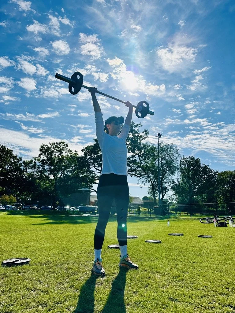 Outdoor fitness in Cranleigh. A woman wearing workout clothes and a cap lifting a barbell overhead on a sunny day at a park with grass, trees, and a bicycle in the background.