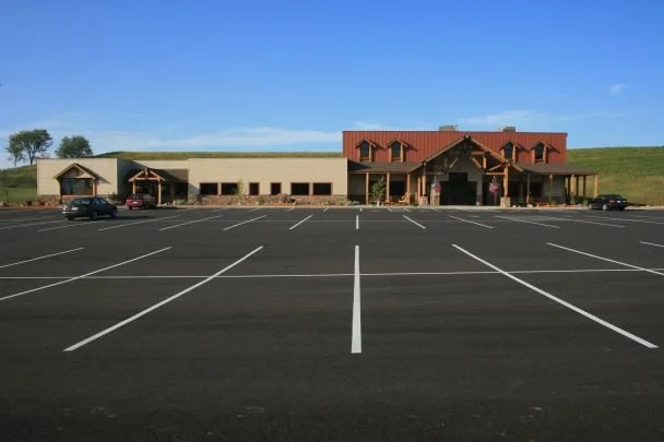 Empty parking lot in front of a large building with a brick facade and a red roof, situated on a grassy hill under a clear blue sky.