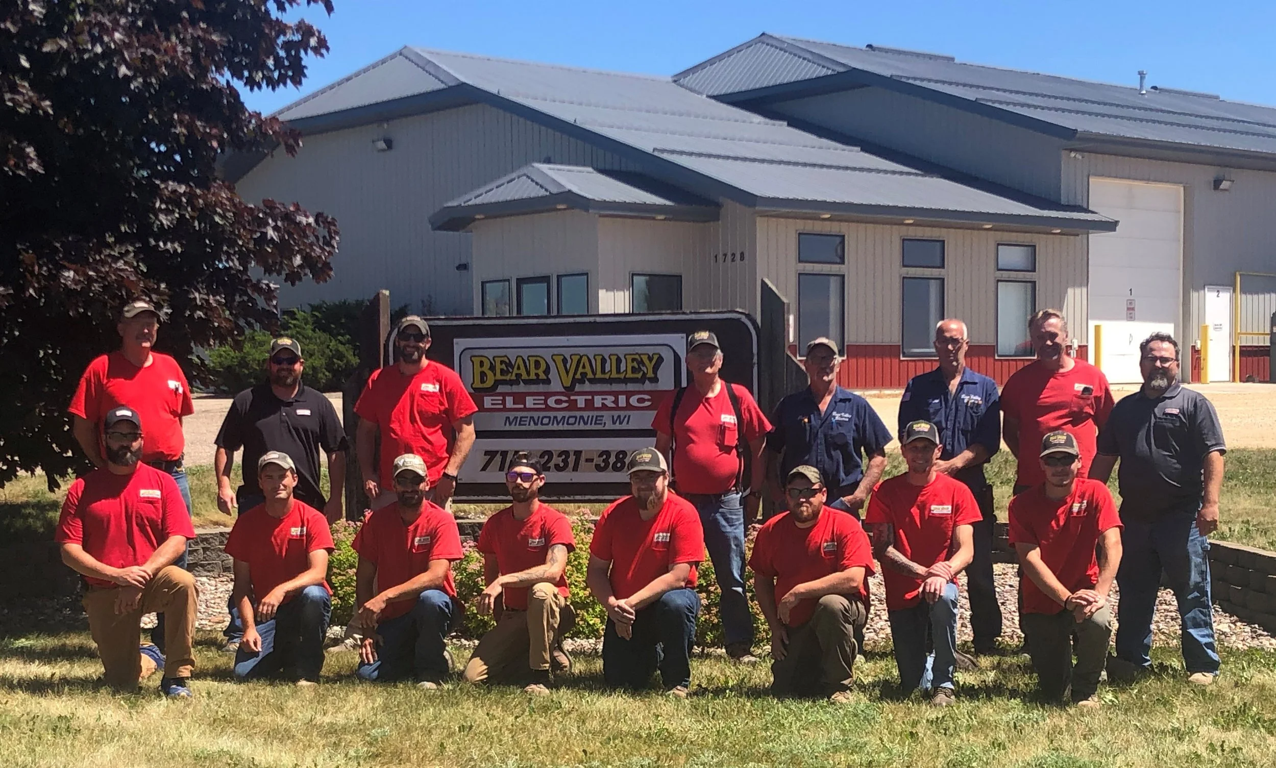 Group of people, some in red shirts and some in blue shirts, posing outside a building with a sign that reads "Bear Valley Electric" in Menomonie, Wisconsin.