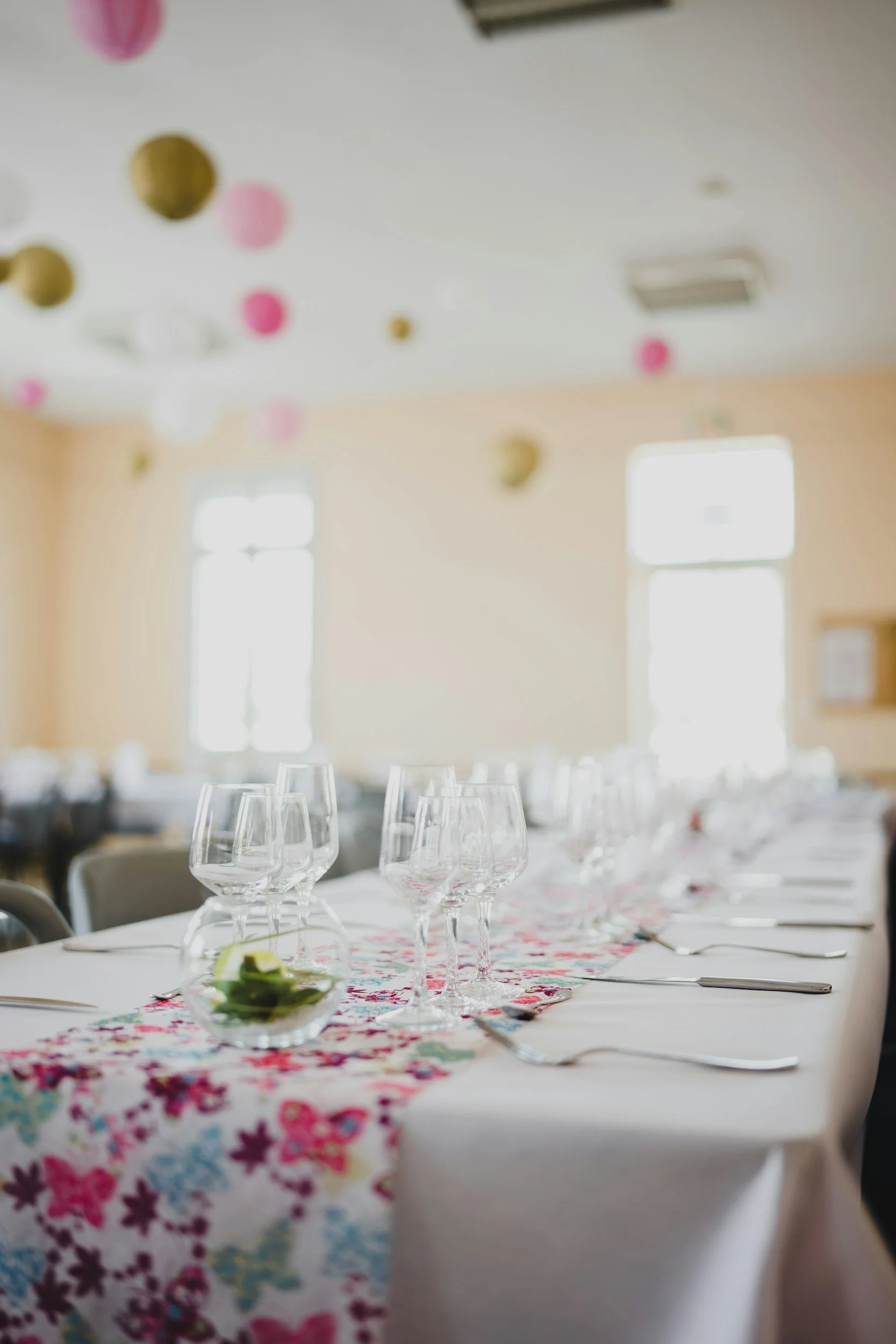 A decorated dining table with wine glasses, cutlery, and a floral table runner, set in a brightly lit room with large windows and hanging paper decorations.
