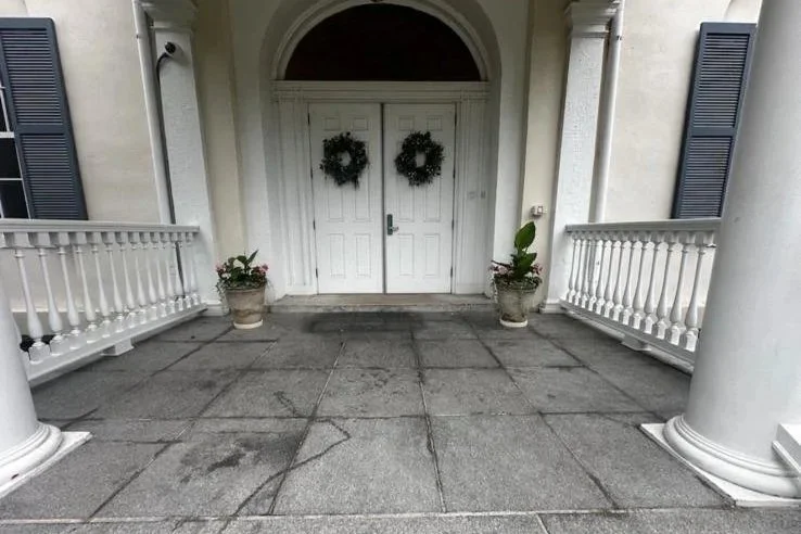 Before photo of Front porch with white double doors adorned with two green wreaths, flanked by potted plants with flowers, and white columns supporting a porch roof.