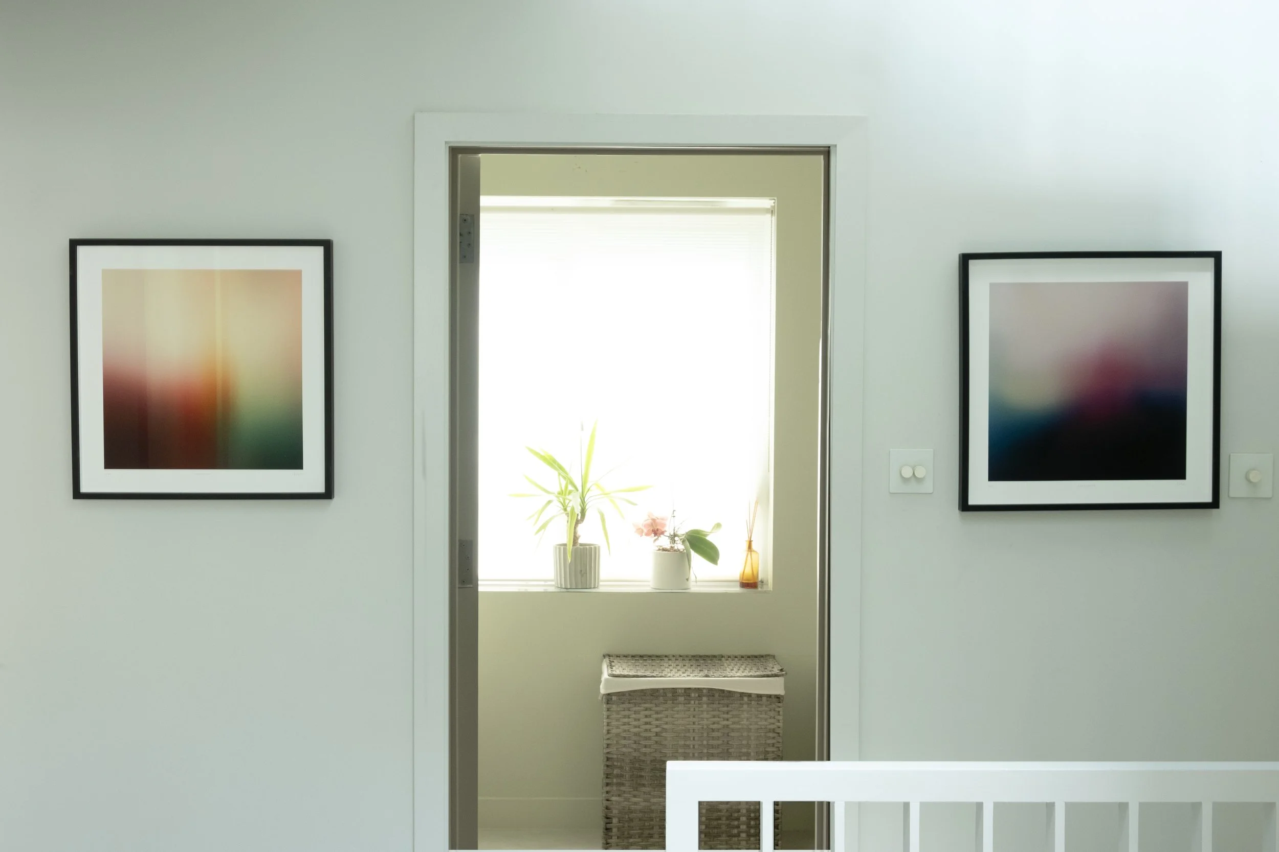 Interior view of a room with a door opening to a sunlit space, decorated with framed abstract art on the walls, a potted plant on the window sill, and a wicker basket below the window.