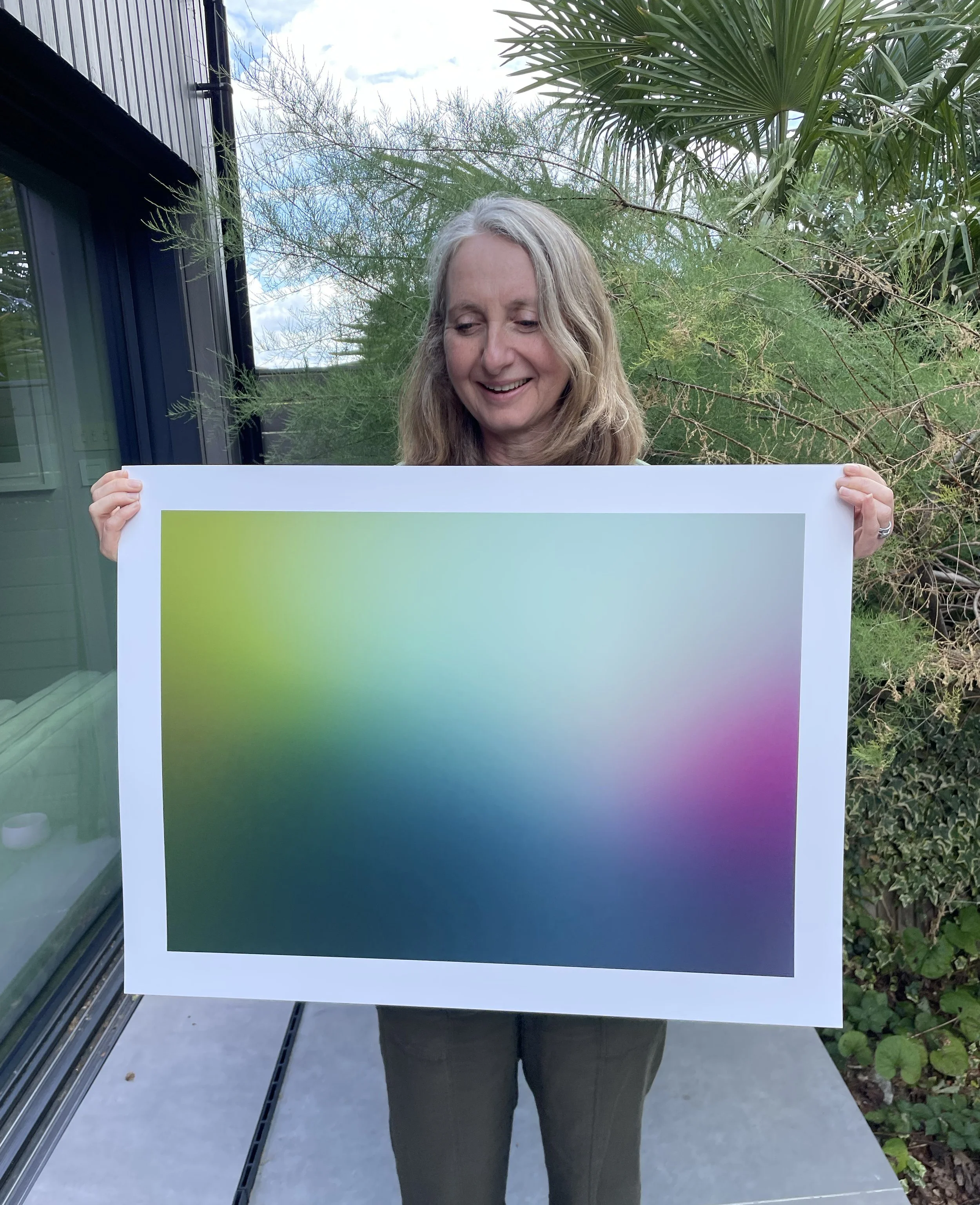 Woman with long blonde hair holding a large white and pastel gradient sign outdoors, with trees and a cloudy sky in the background.