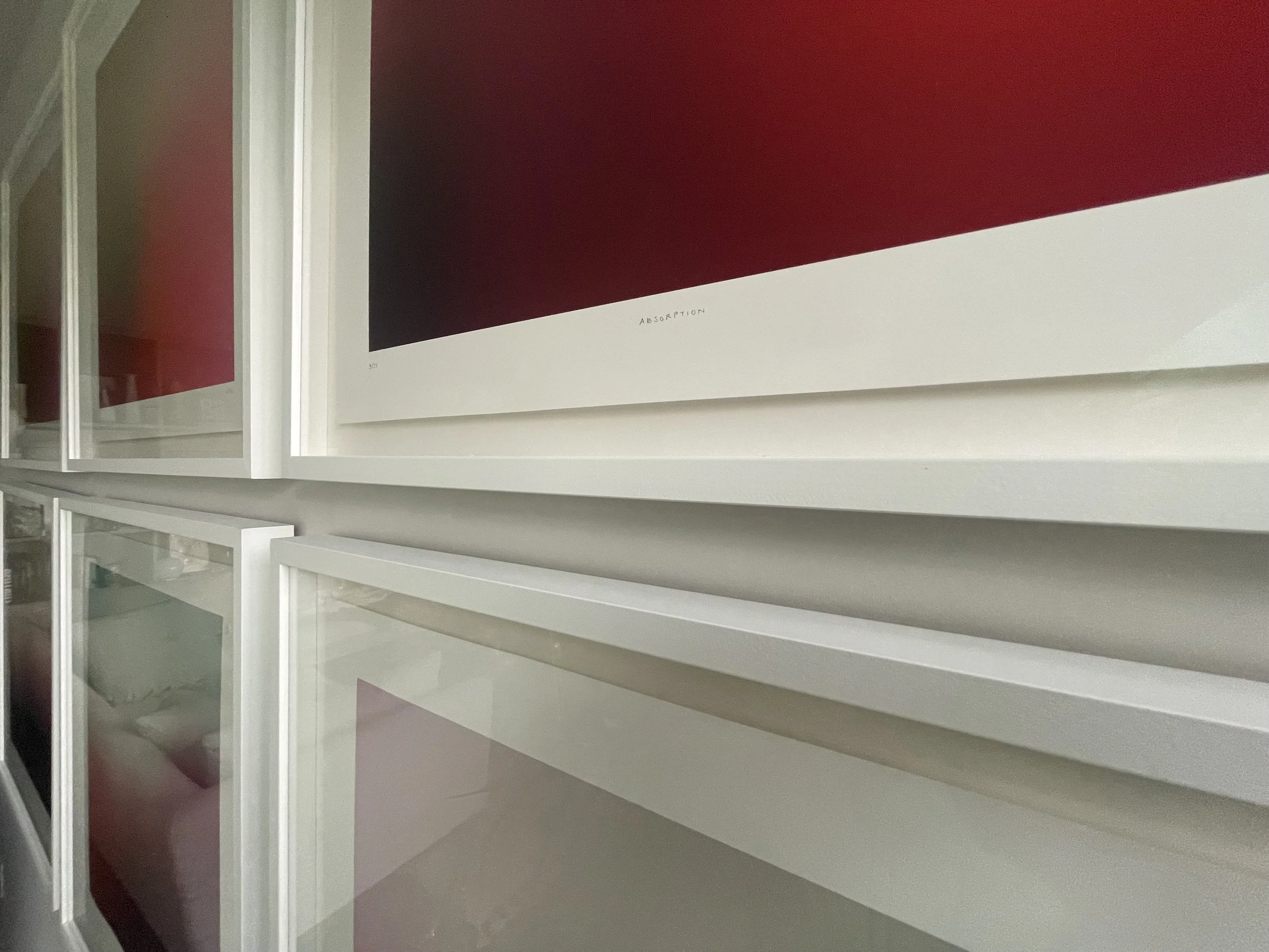 A modern interior wall with white  large framed artworks, one titled 'Absorption' featuring a gradient of black to red, mounted above the shelves.