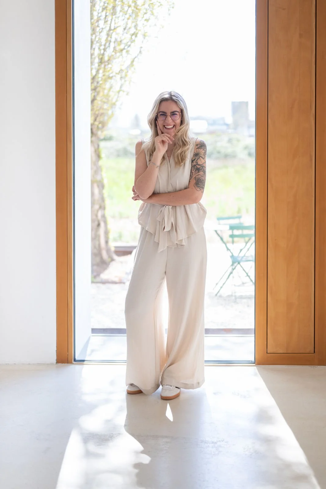 Eine lachende junge Frau mit Brille steht in einem offenen Eingang zwischen Holzrahmen. Sie trägt ein beiges, lässiges Outfit und hat Tattoos am Arm.