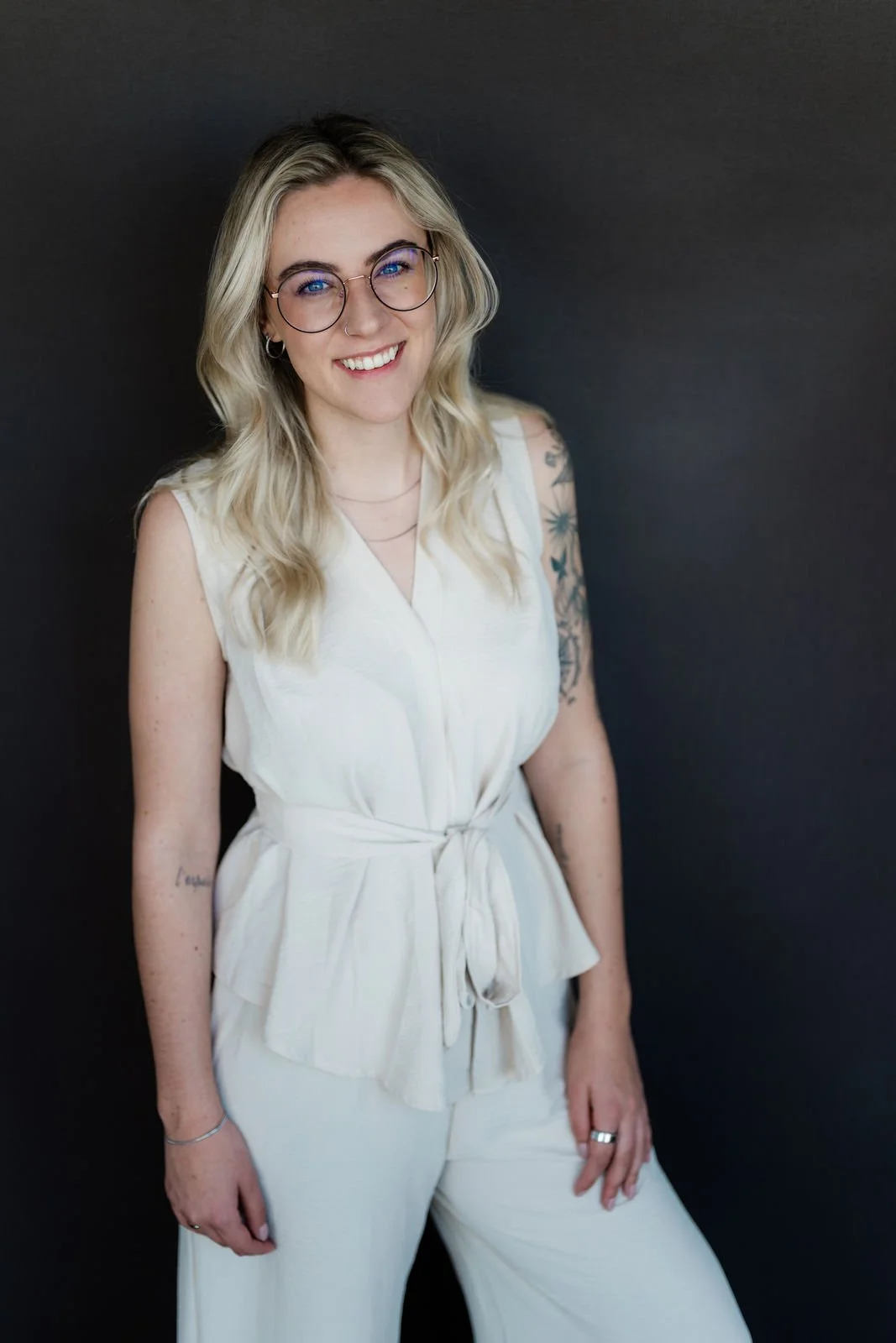 Junge Frau mit Brille, langen, blonden Haaren und Tattoos, trägt weiße Kleidung, lächelt vor dunkelgrauem Hintergrund.