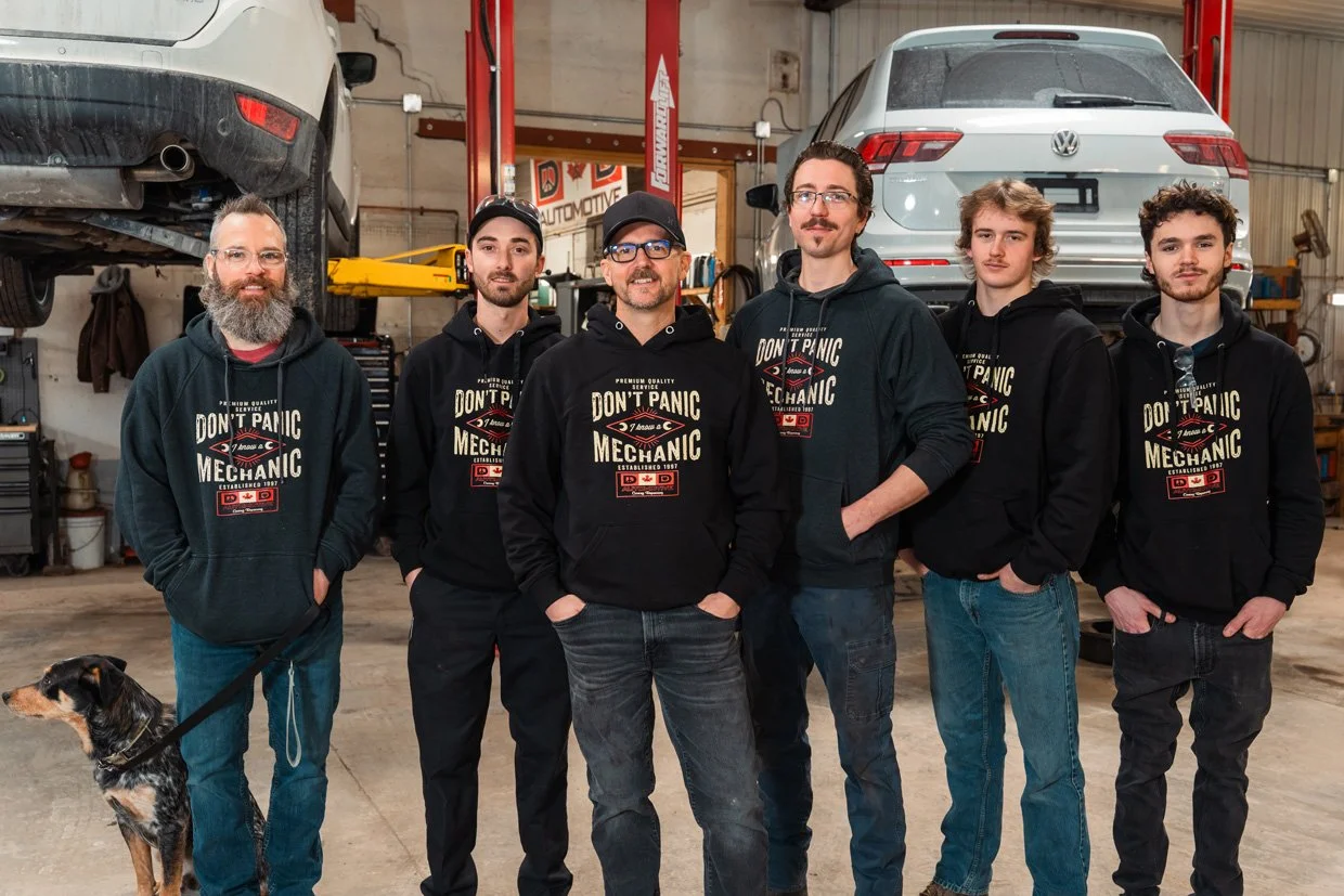 Six men wearing black hoodies with 'Don't Panic Mechanic' logo standing in a car repair garage with two elevated vehicles and a dog on a leash.