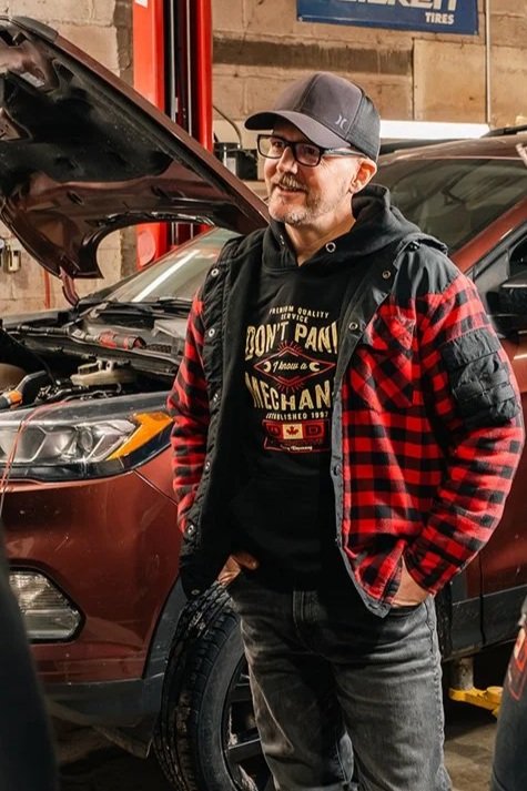 A man with glasses, a beard, and a cap is standing next to a car with its hood open in a garage. He is wearing a black hoodie and a red and black plaid jacket.