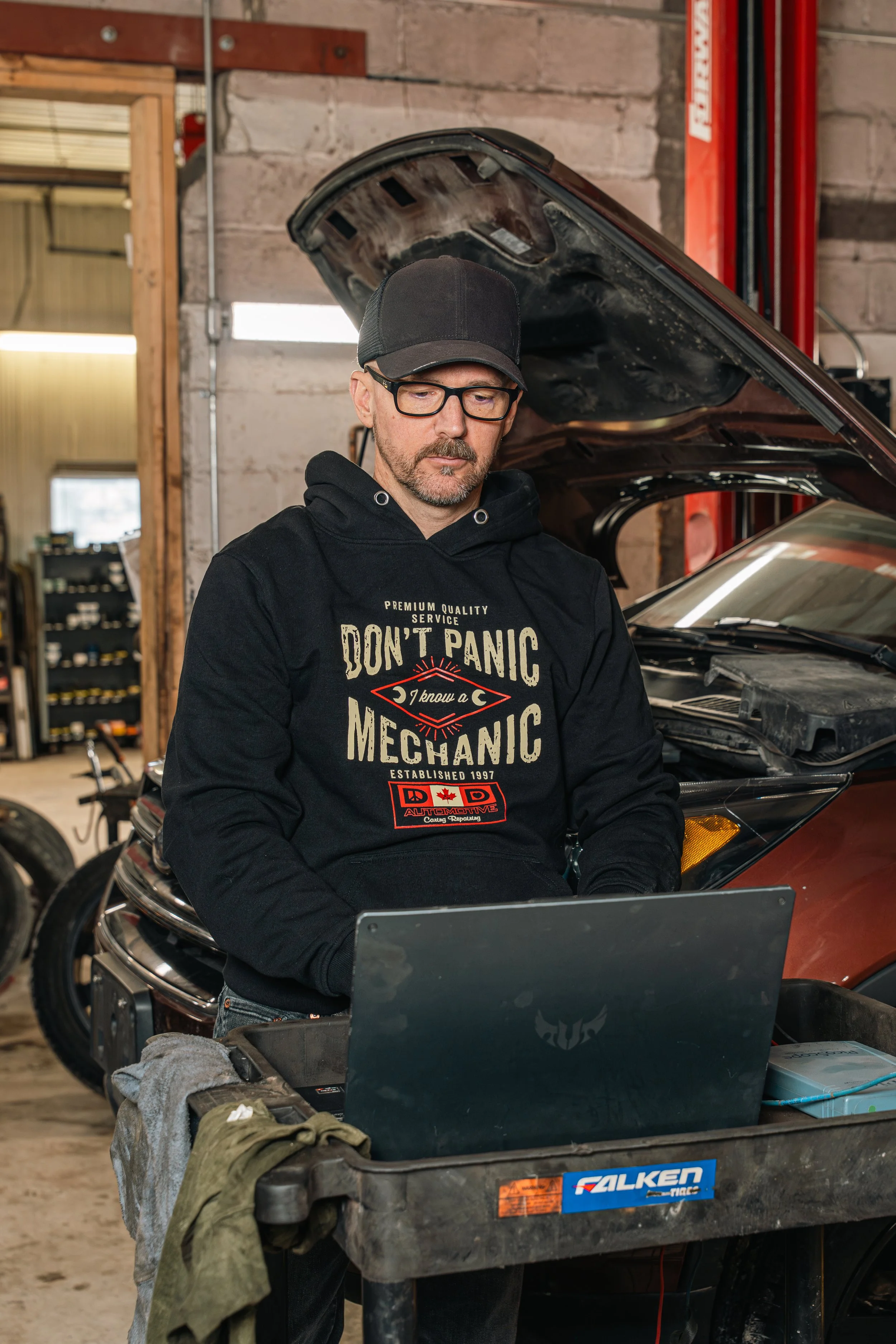 A man wearing glasses, a black cap, and a black hoodie stands in a garage next to a car with its hood open, working on a laptop placed on a workbench.