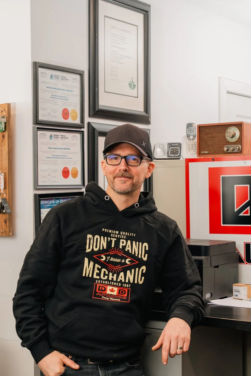 A man wearing glasses, a black cap, and a black hoodie with a mechanic quote, standing in an office or workshop with framed certificates on the wall and a red and black signs.