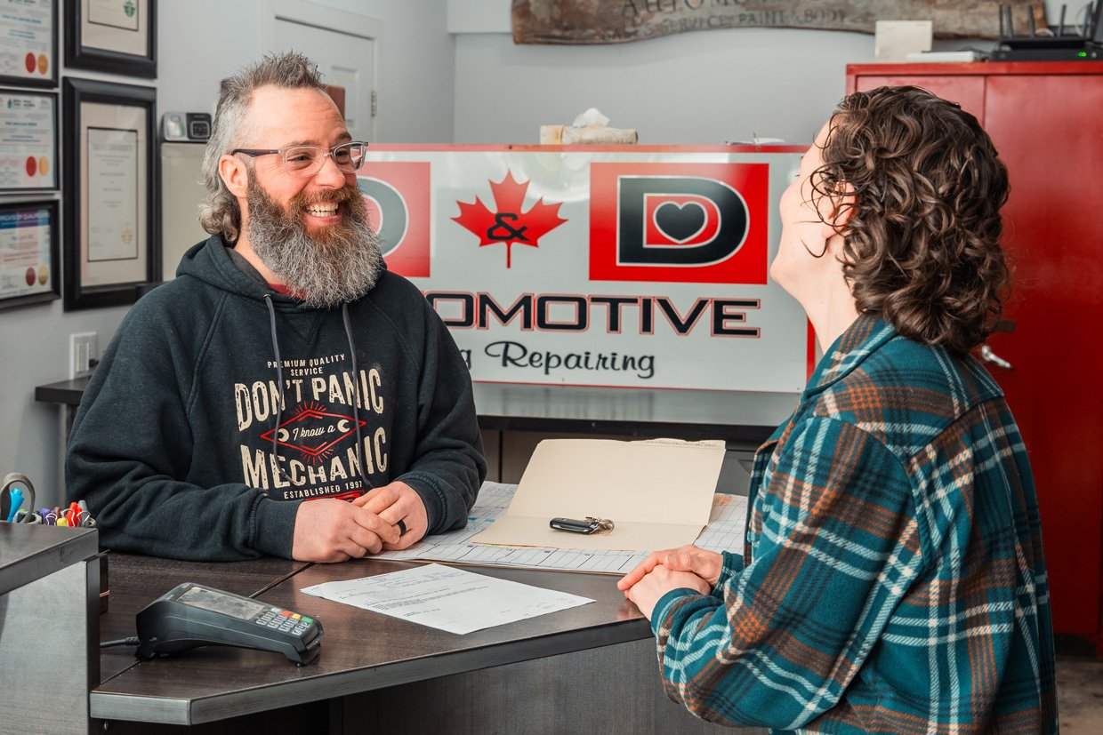 A man with glasses and a beard smiling and talking to a woman with curly hair at a reception desk in an automotive repair shop.