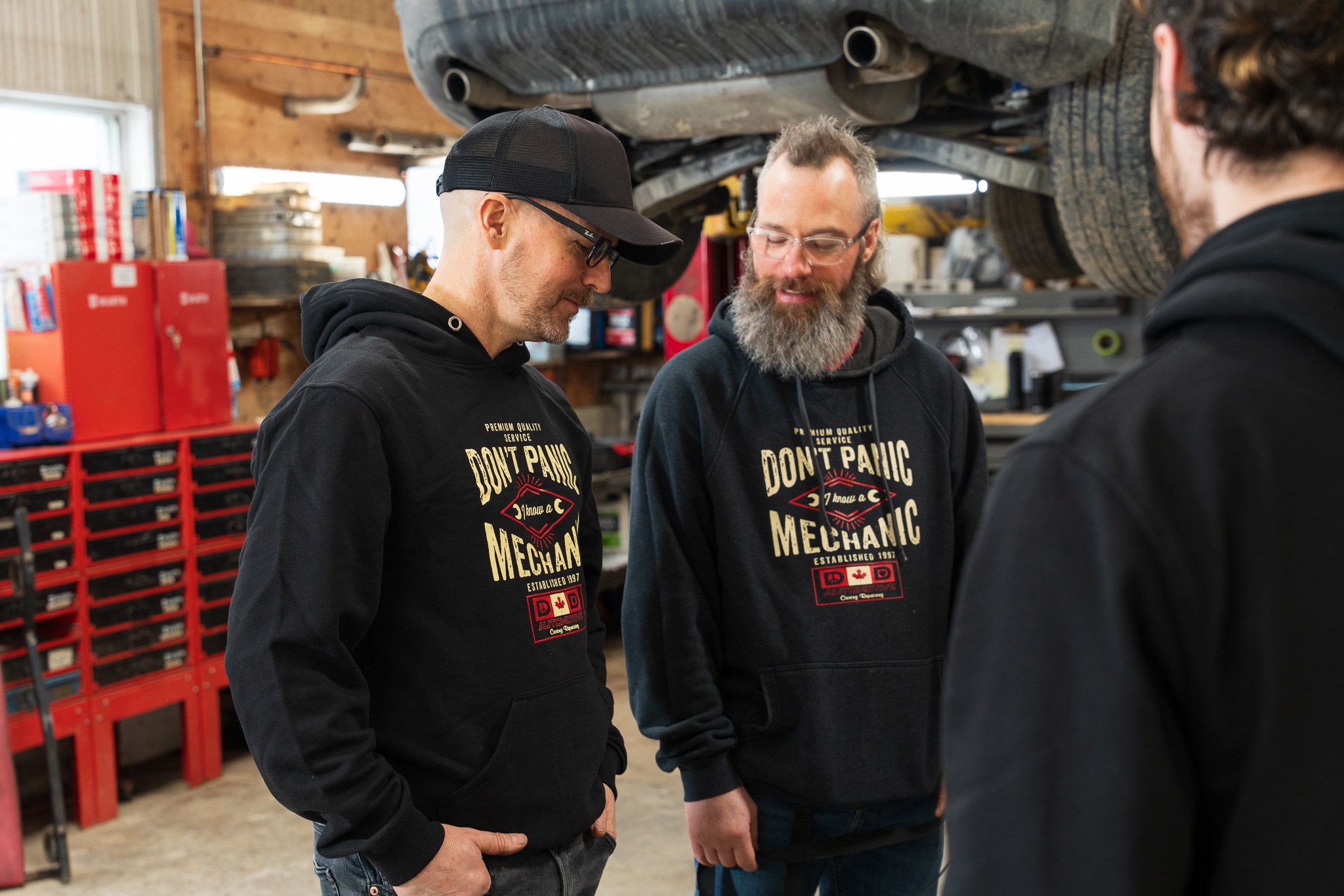 Three men in a garage or workshop, wearing black hoodies with the text 'Don't Panic Mechanic' and a wrench graphic, engaged in conversation under a vehicle lifted above them.