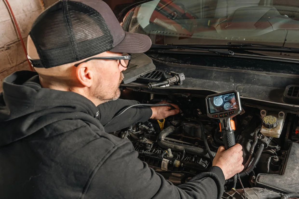 A man wearing a black hoodie, glasses, and a trucker hat inspecting a car engine using an electronic diagnostic tool in a garage.