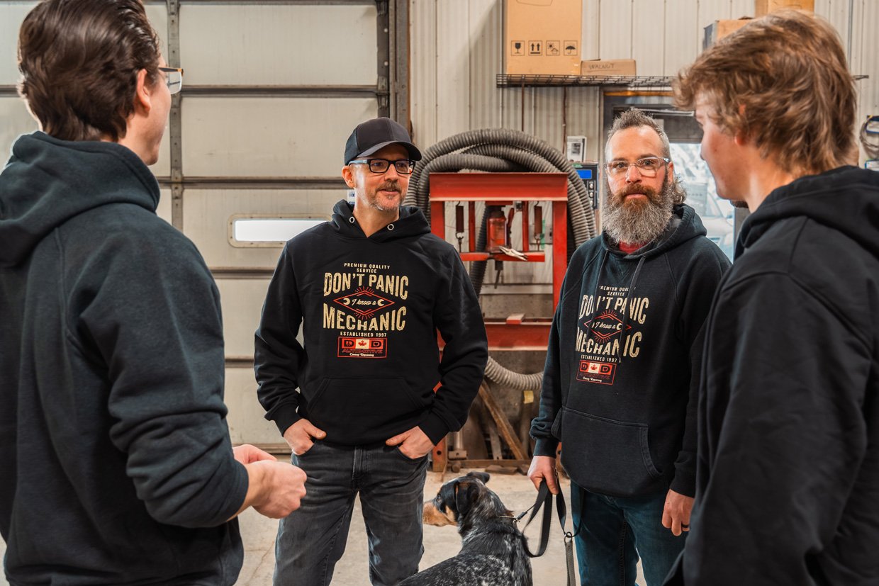 Four men talking inside a garage or workshop with a dog. All four are wearing black hoodies with the text "Don't Panic Mechanic." The men are casually dressed, and two of them have beards and glasses. One man on the right has a leash with a dog, which is partially visible.