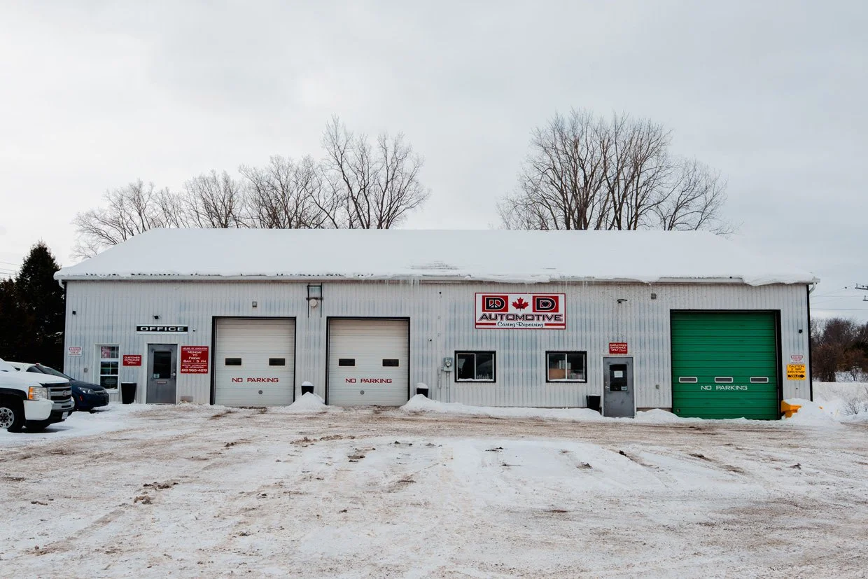 A winter scene of an automotive repair shop with snow-covered ground. The building has a white exterior with multiple garage doors, including a large green door on the right. Signs indicate no parking areas and the shop's sign reads DD Automotive with a red maple leaf. Several vehicles are parked outside.