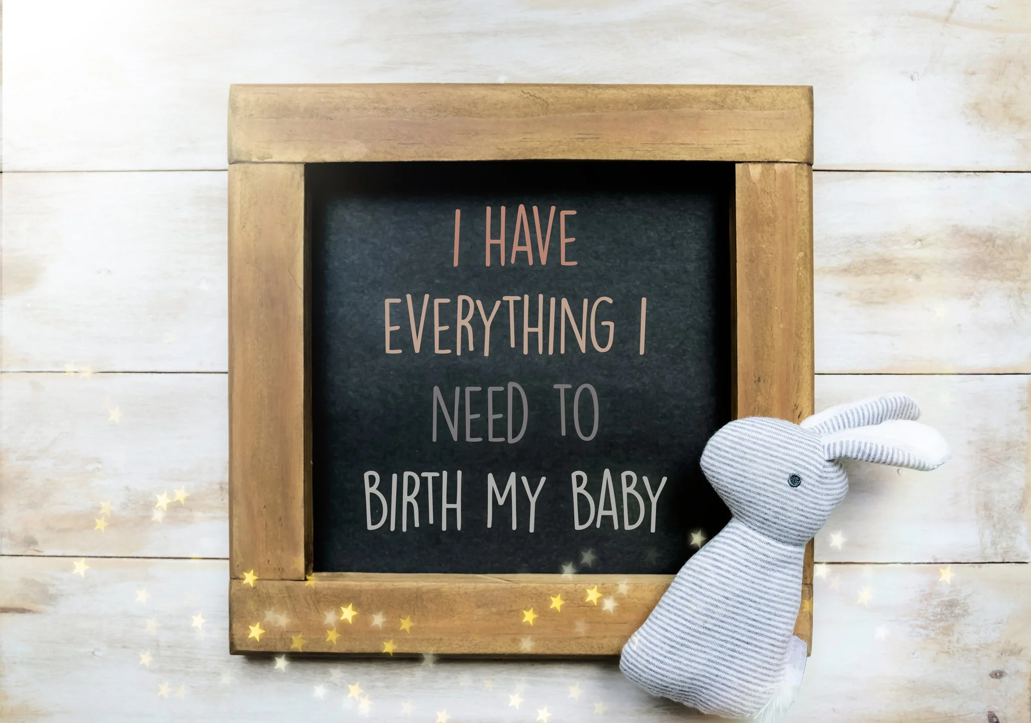 A blackboard with a wooden frame displaying the message 'I have everything I need to birth my baby.' Sitting next to the blackboard is a gray striped plush bunny toy. The background is a white wooden surface with small gold star decorations.