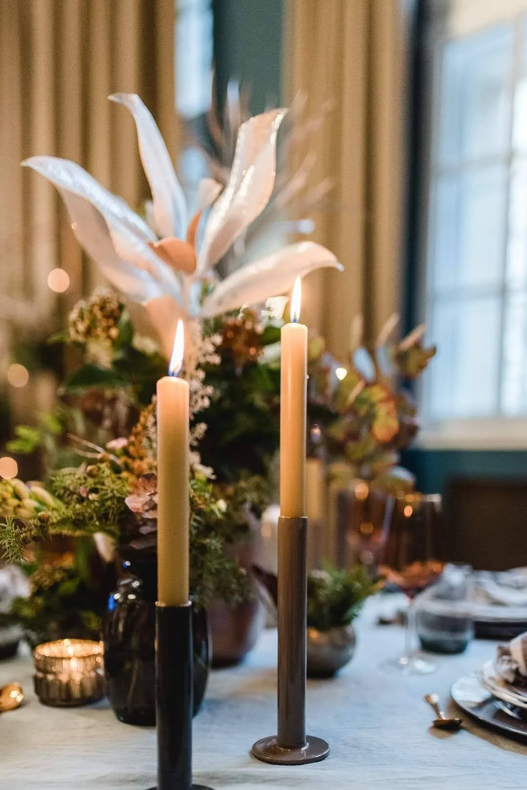 Ein festlich gedeckter Tisch mit Kerzen, Blumen und Weingläser in einem Raum mit Fenster und Vorhängen.