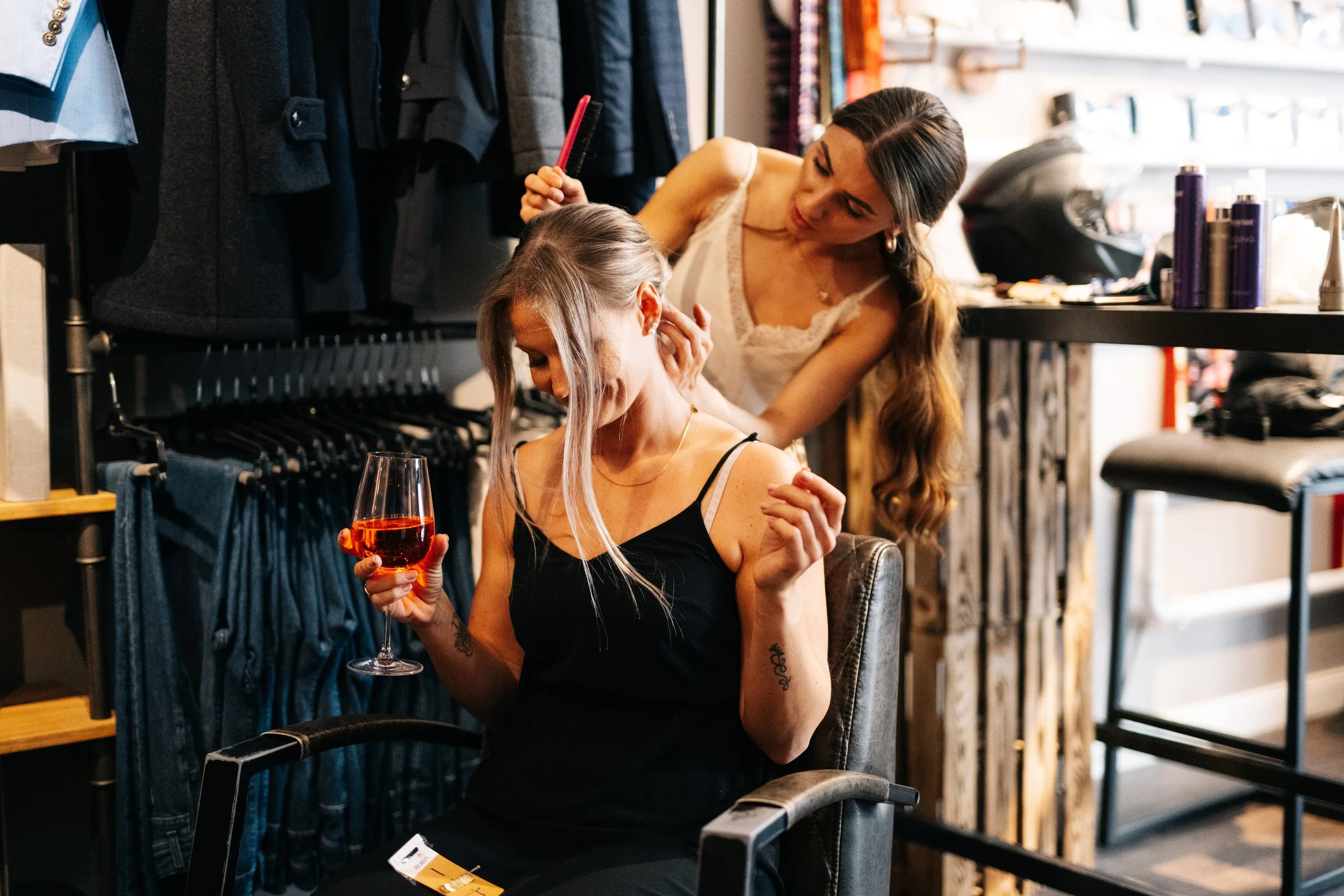 Zwei Frauen in einem Friseursalon, eine sitzt im Friseurstuhl mit einem Glas Rotwein in der Hand, die andere stylt ihre Haare.