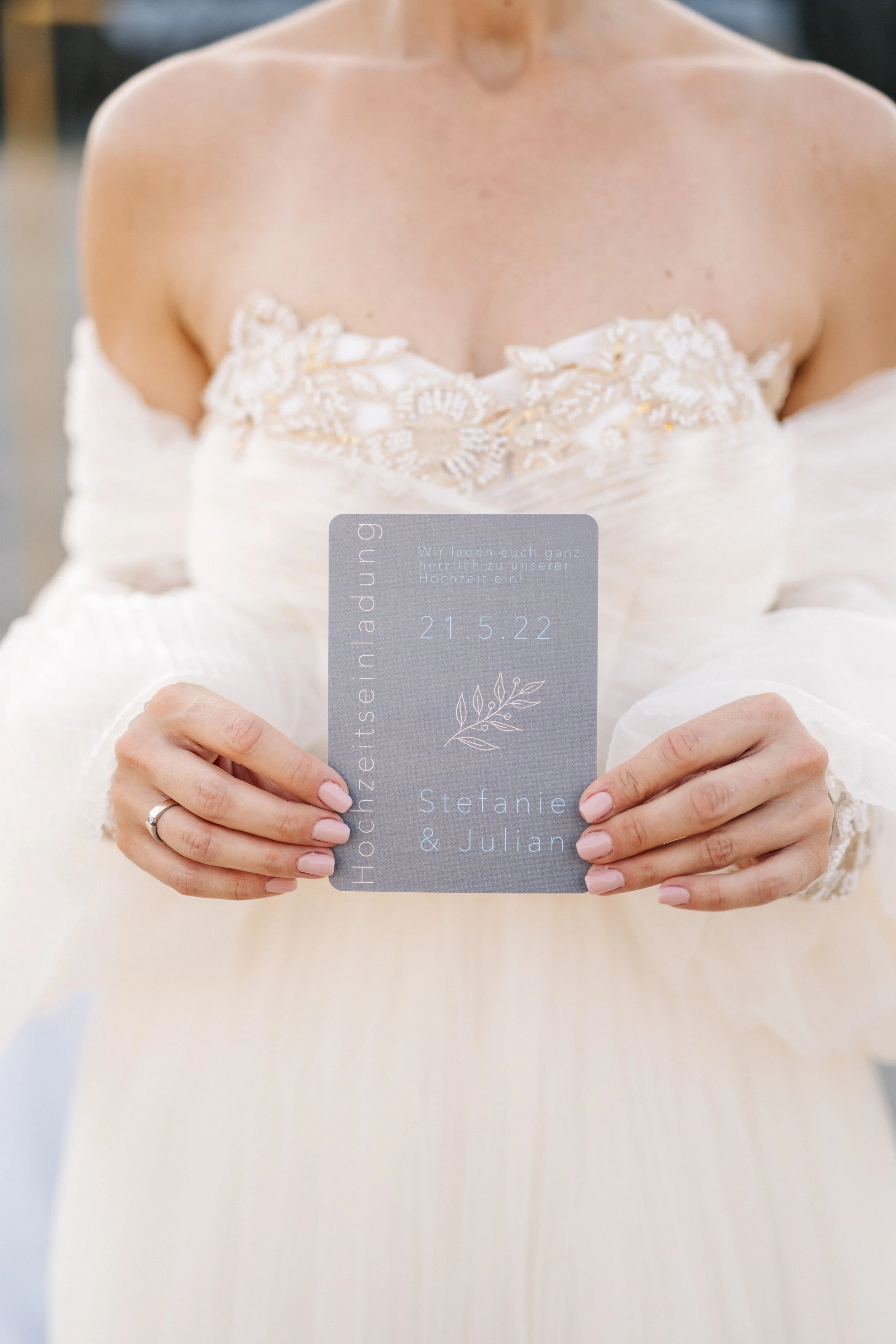 Braut in weißem Hochzeitskleid hält Einladungskarte für Hochzeit mit Namen und Datum.