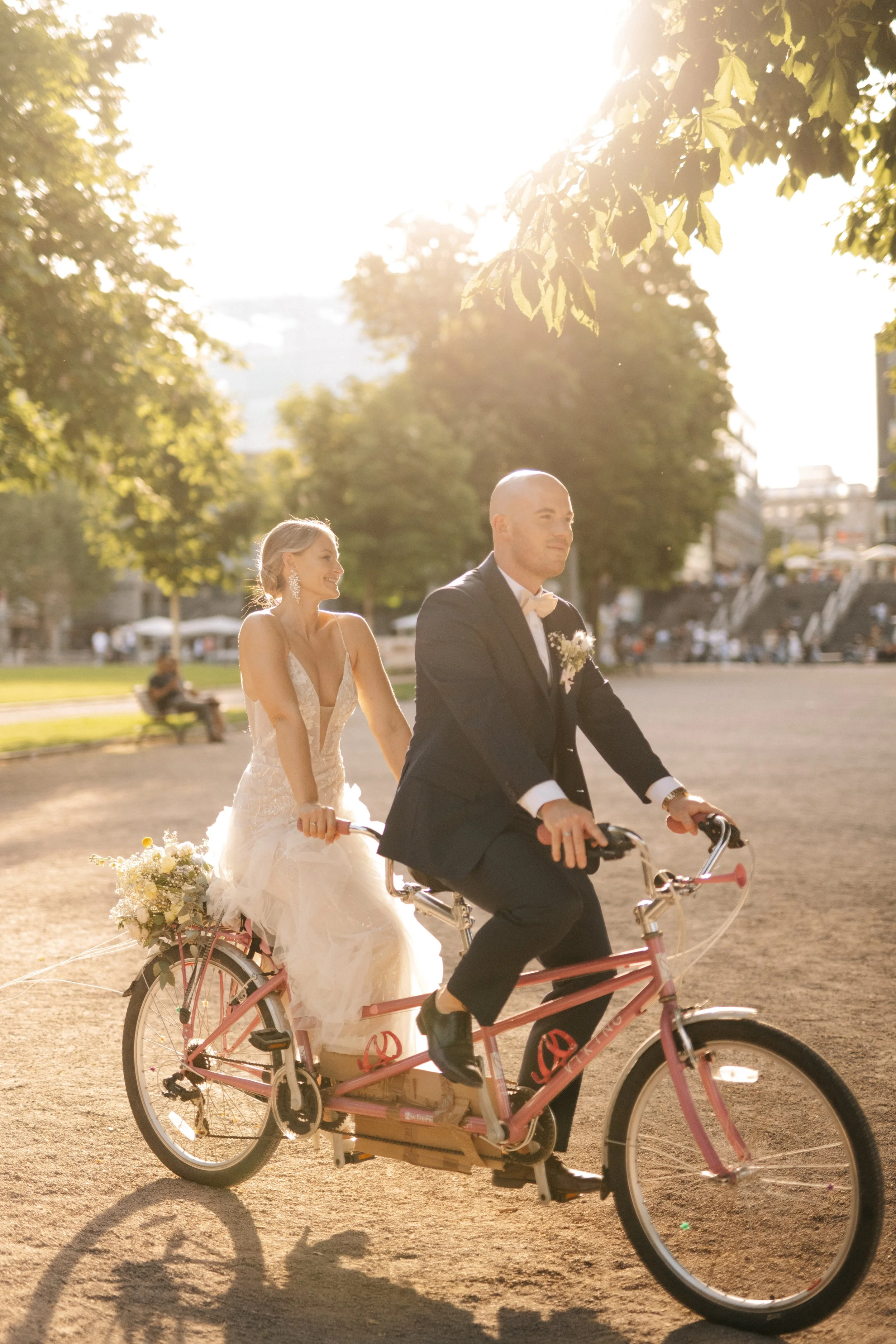 Ein Brautpaar auf einem Fahrrad im Park bei Sonnenuntergang, die Braut trägt ein weißes Abendkleid, der Bräutigam einen schwarzen Anzug.