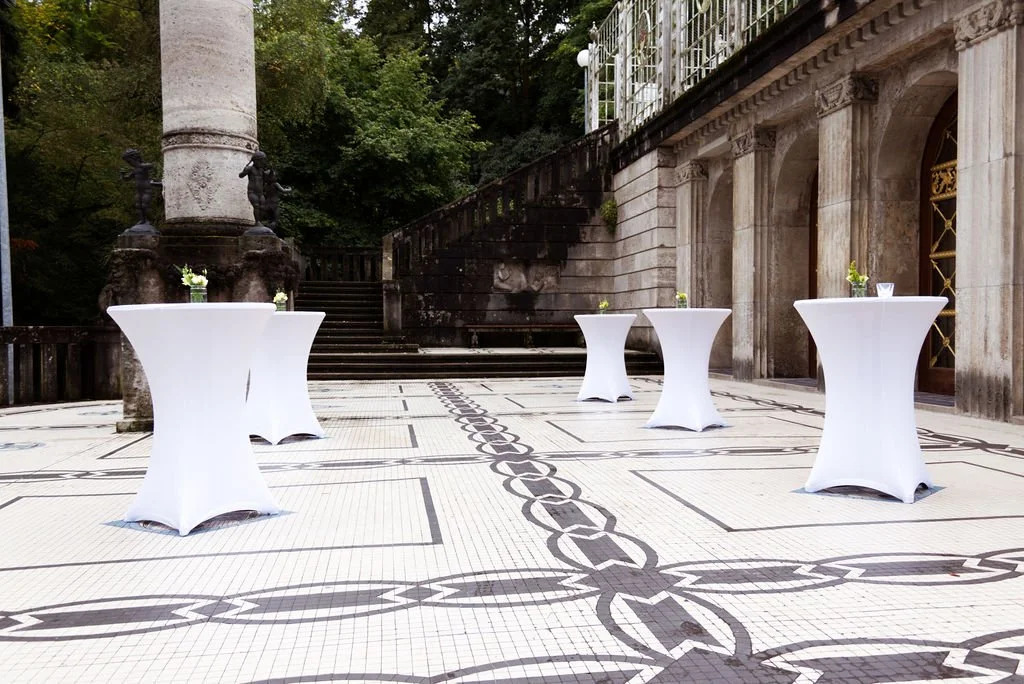 Vier Stehtische mit weißen Hussen und kleinen Blumenvasen, auf einer eleganten, gepflegten Terrasse im Freien, umgeben von antiker Architektur und grünen Bäumen.