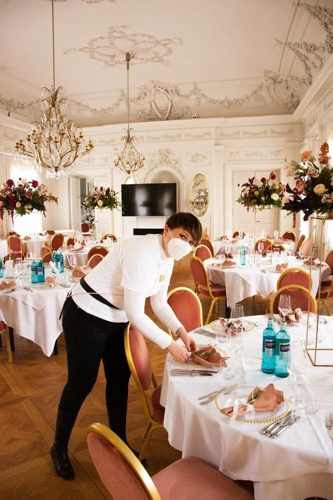 Eine Person in einem festlich gedeckten Saal richtet den Tisch für eine Veranstaltung. Die Person trägt eine Schutzmaske, ein weißes Hemd und schwarze Hose. Der Saal ist elegant mit Blumenarrangements, Kronleuchtern und festlicher Dekoration eingeric