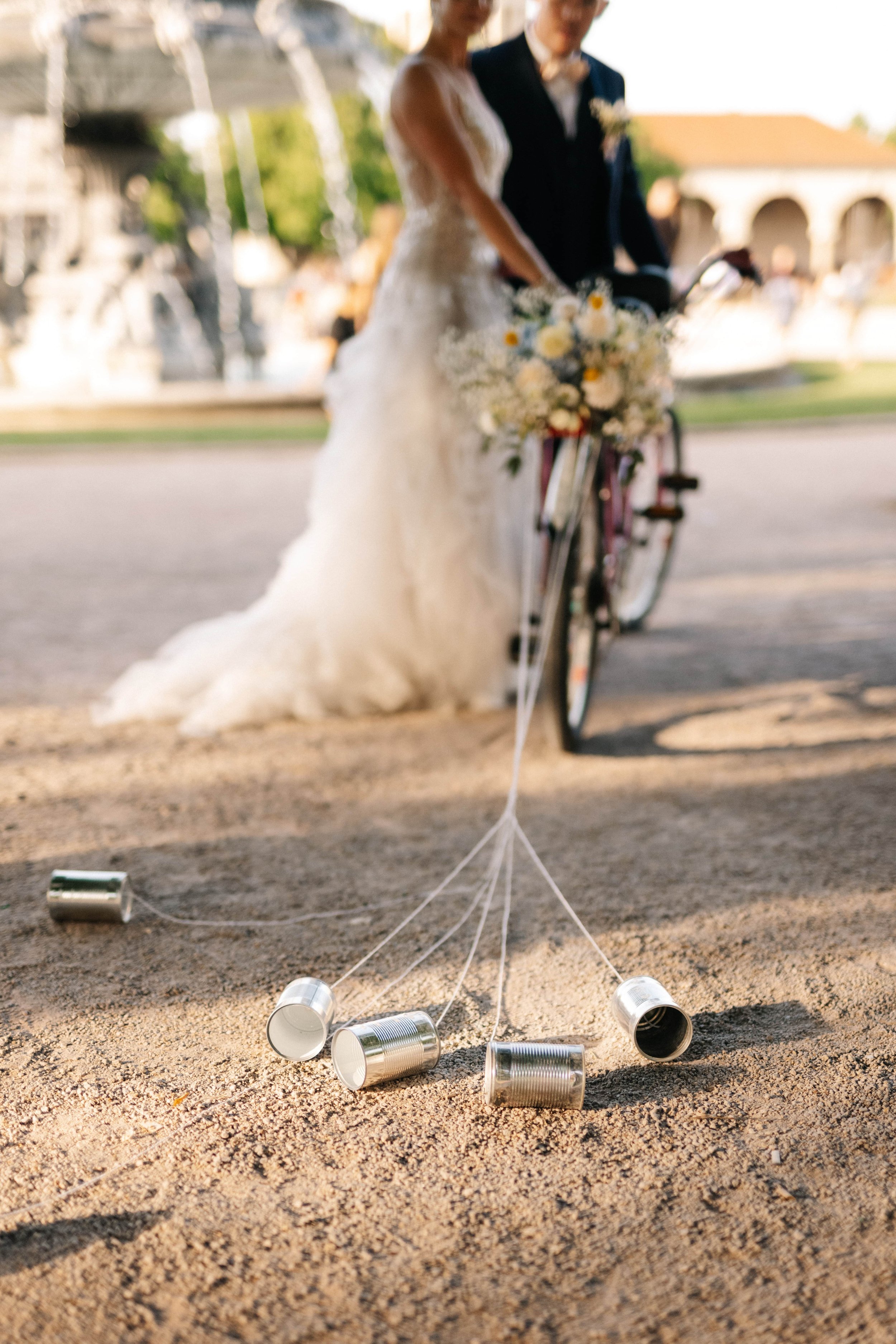 Ein Brautpaar auf einem Fahrrad, das mit Blumen geschmückt ist, steht auf einem Sandweg. Vor ihnen liegen fünf leere Konservendosen, die an Schnüren befestigt sind.
