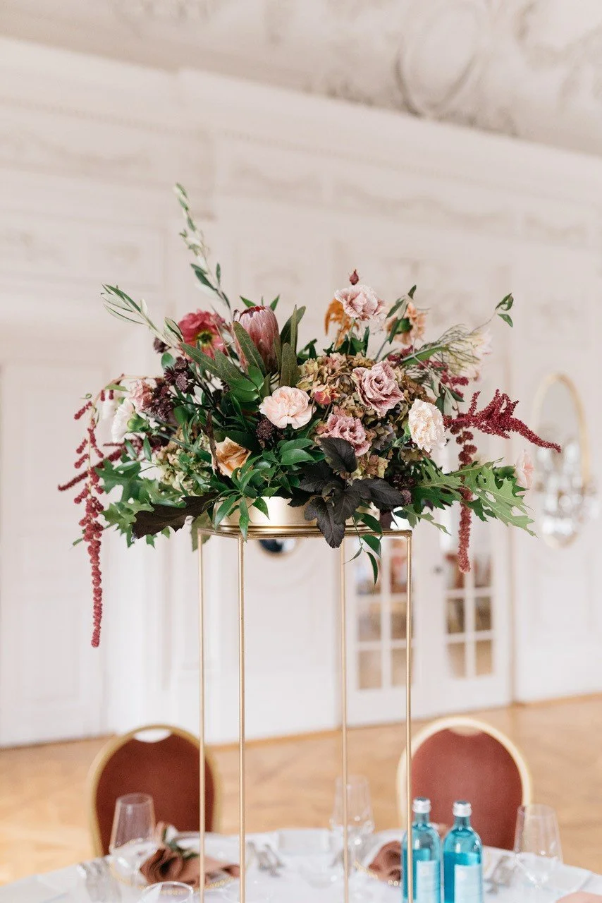 Blumenarrangement auf goldfarbenem Stehtisch in einem festlich gedeckten Raum.