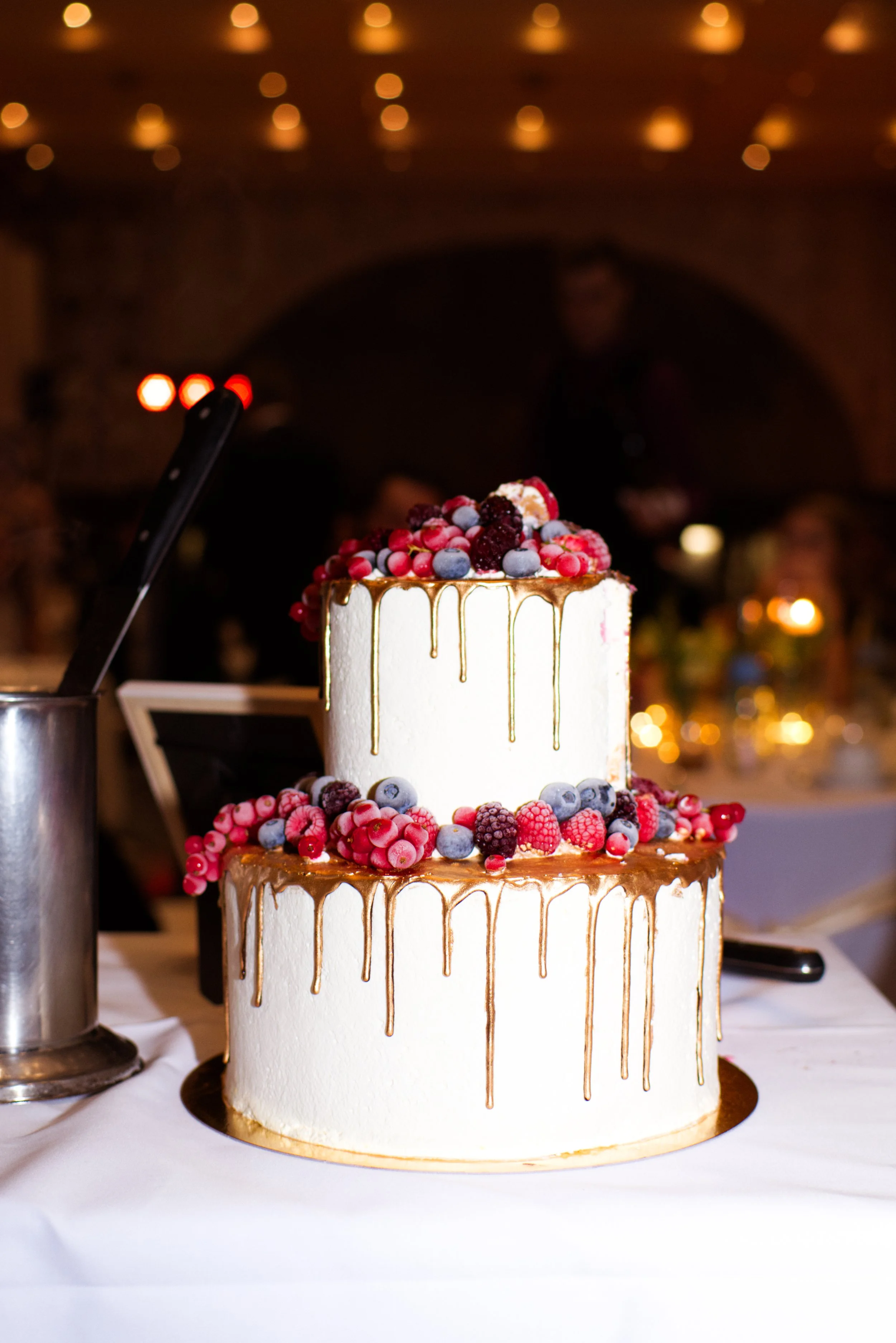 Zweistöckige Hochzeitstorte mit weißen Zuckerguss, verziert mit buntem Beerenobst und einem goldenen Drip-Glanz.