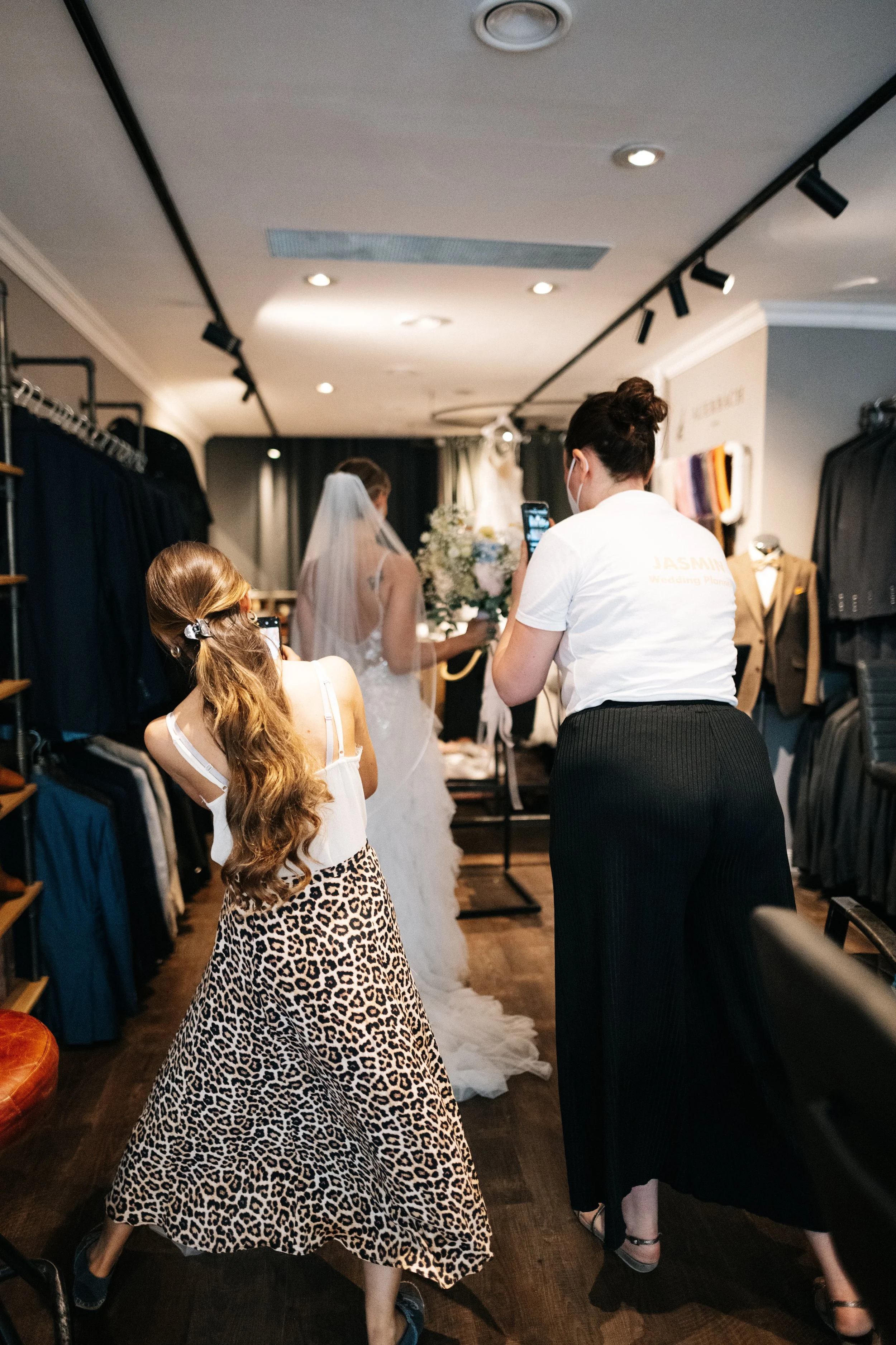 Drei Frauen im Modegeschäft, eine there auf einen Brautkleid, andere zwei fotografieren oder filmen es. Eine Frau trägt eine Leopardenschwanzrock, die andere trägt eine weiße T-Shirt mit Text. Im Hintergrund eine Braut im weißen Kleid, eine Blumenstr