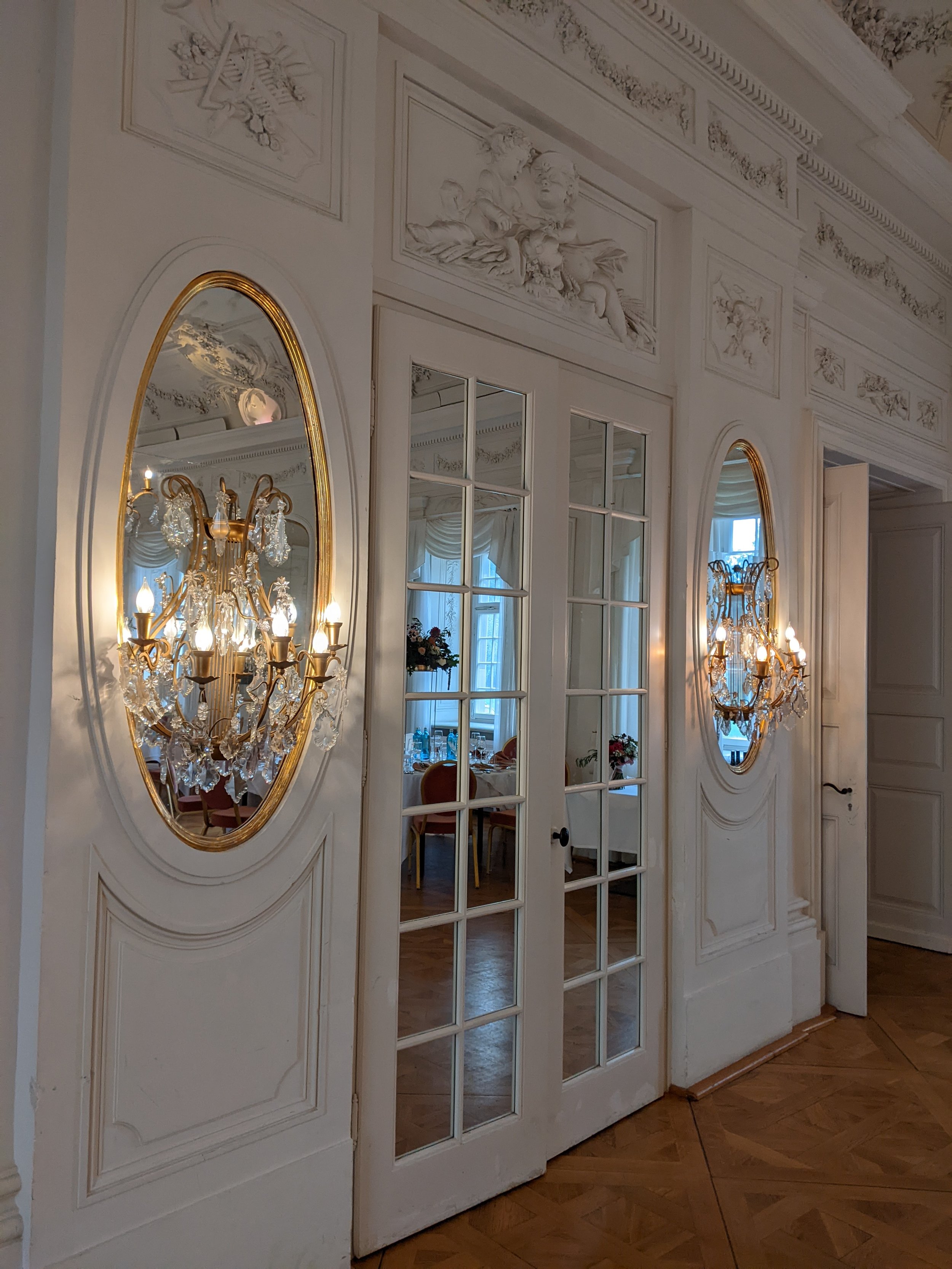 Elegantes, weißes Rauminnere mit goldgerahmten Spiegeln und Kronleuchtern, die in den Spiegeln reflektiert werden. Deko-Details und Fenster im Hintergrund.