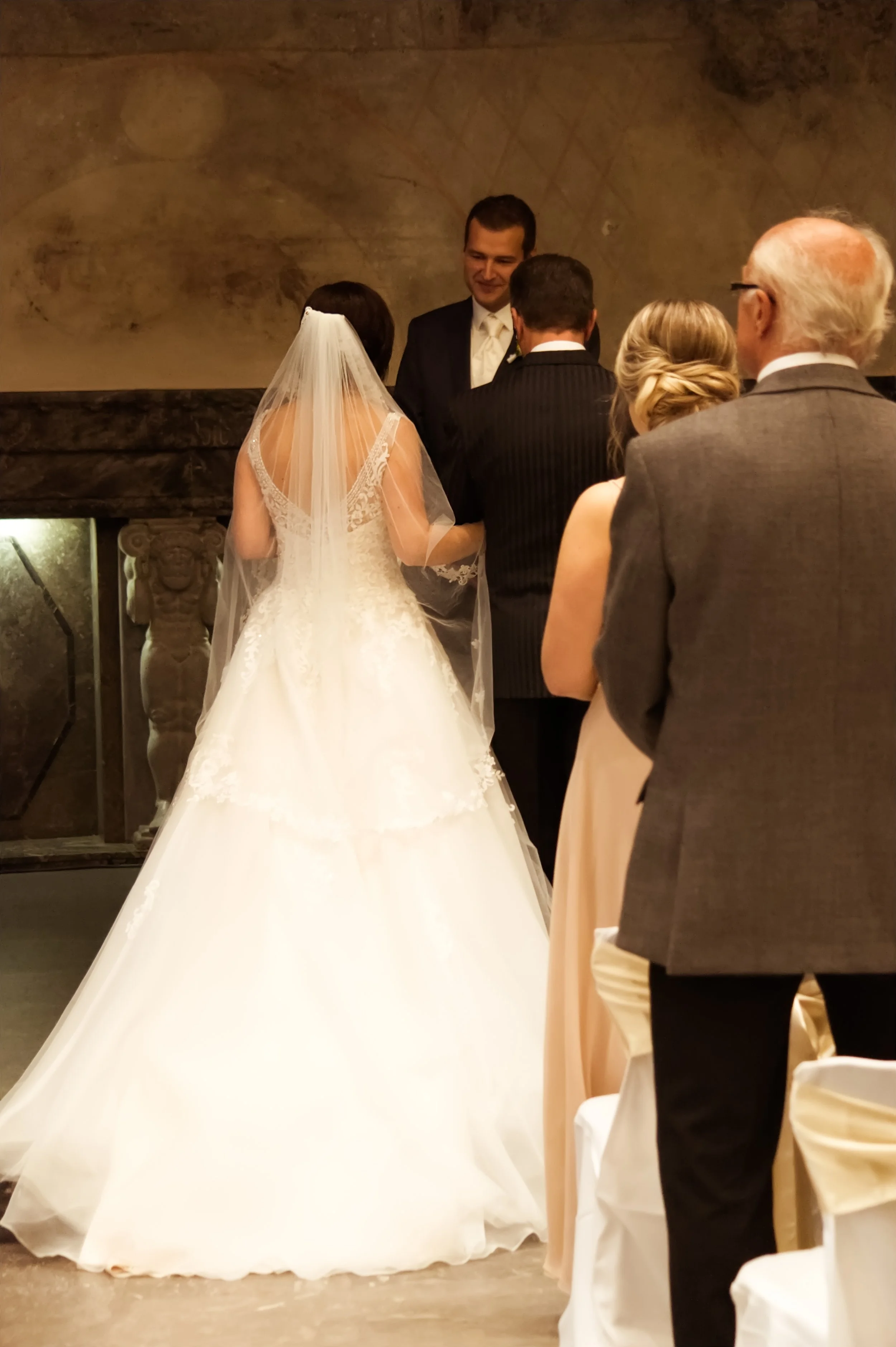 Braut in weißem Kleid und Schleier bei einer Hochzeitszeremonie, umgeben von Gästen in formaler Kleidung, in einem Raum mit dunkler, warm beleuchteter Atmosphäre.