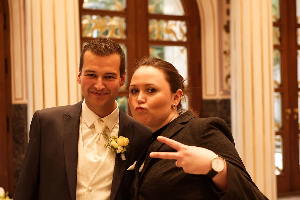 Ein Mann im Anzug und eine Frau in dunkler Kleidung posieren im Raum mit großen Fenstern im Hintergrund. Die Frau zeigt ein Peace-Zeichen mit ihrer Hand.