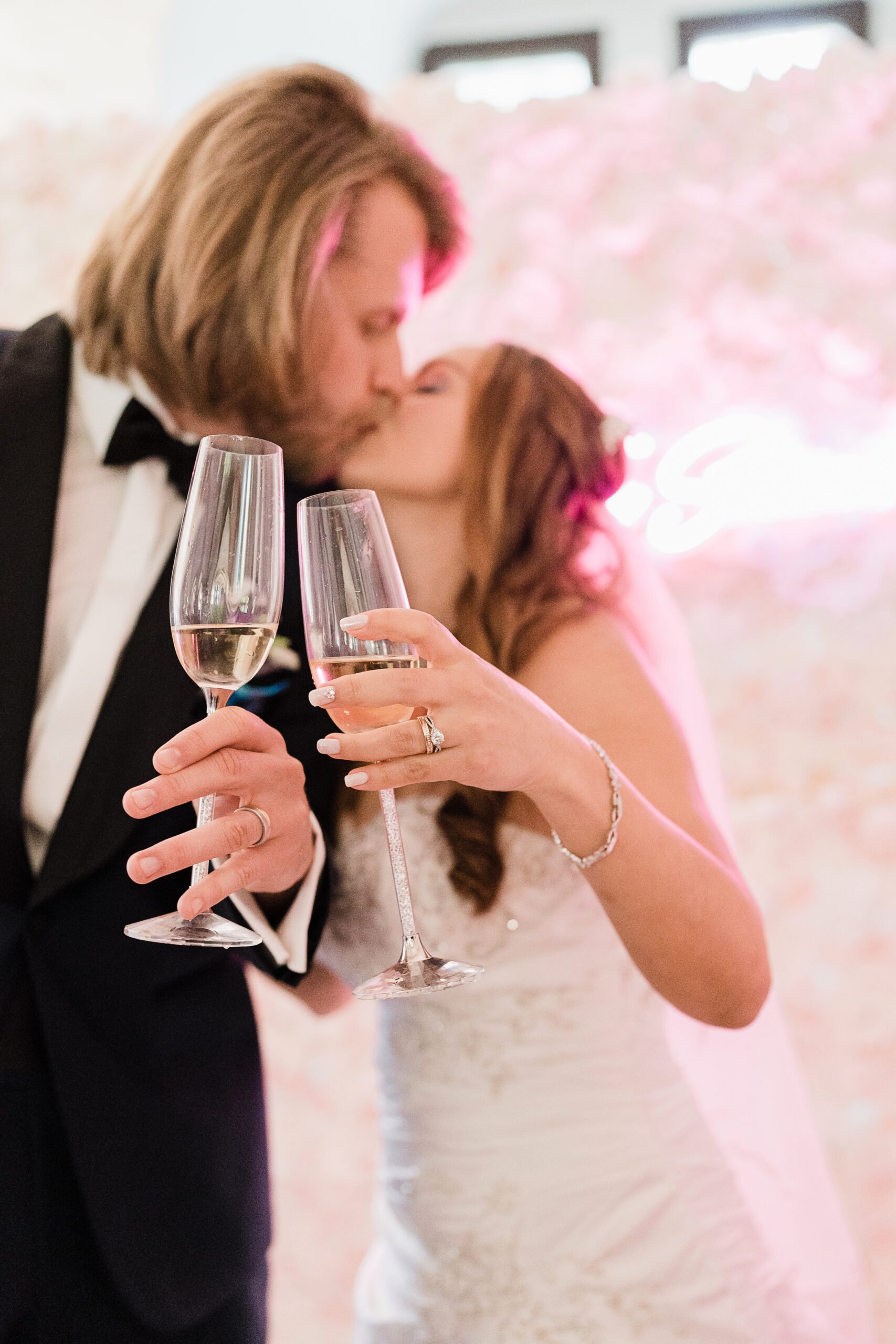 Ein Hochzeitsfoto von einem Paar, das sich küsst, während sie Gläser Champagner halten, im Hintergrund eine rosa Beleuchtung.