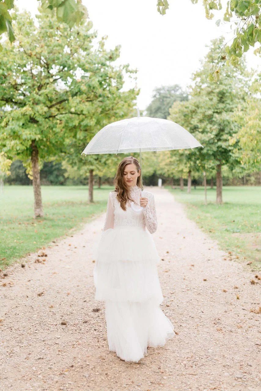 Junge Frau in weißen Hochzeitskleid, die einen transparenten Regenschirm hält, wandert auf einem Weg in einem Park umgeben von grünen Bäumen.