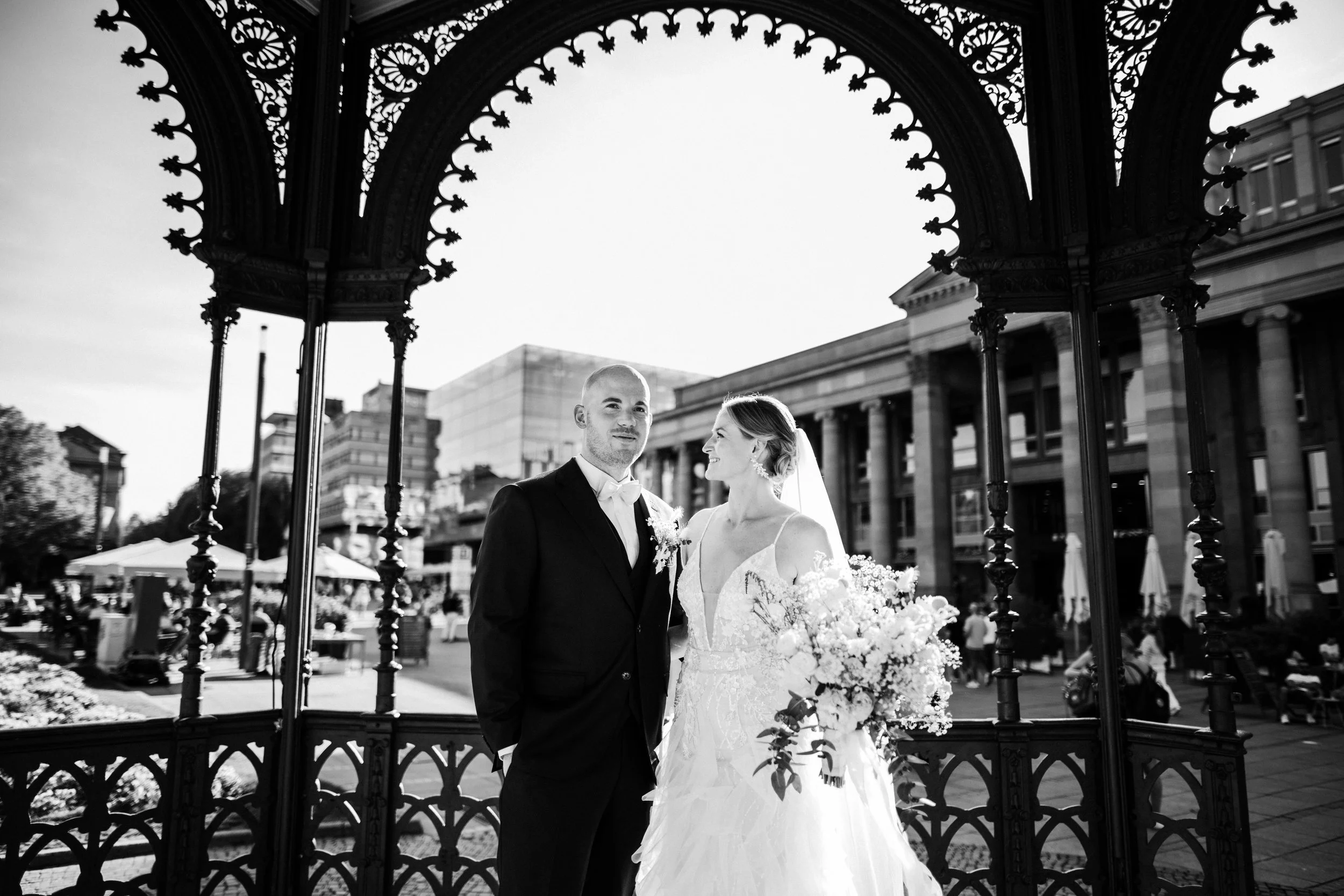 Brautspaare während einer Hochzeit, stehend unter einem dekorativen Pavillon im Freien, im Hintergrund Gebäude und Menschen in einer Stadt, in Schwarz-Weiß.