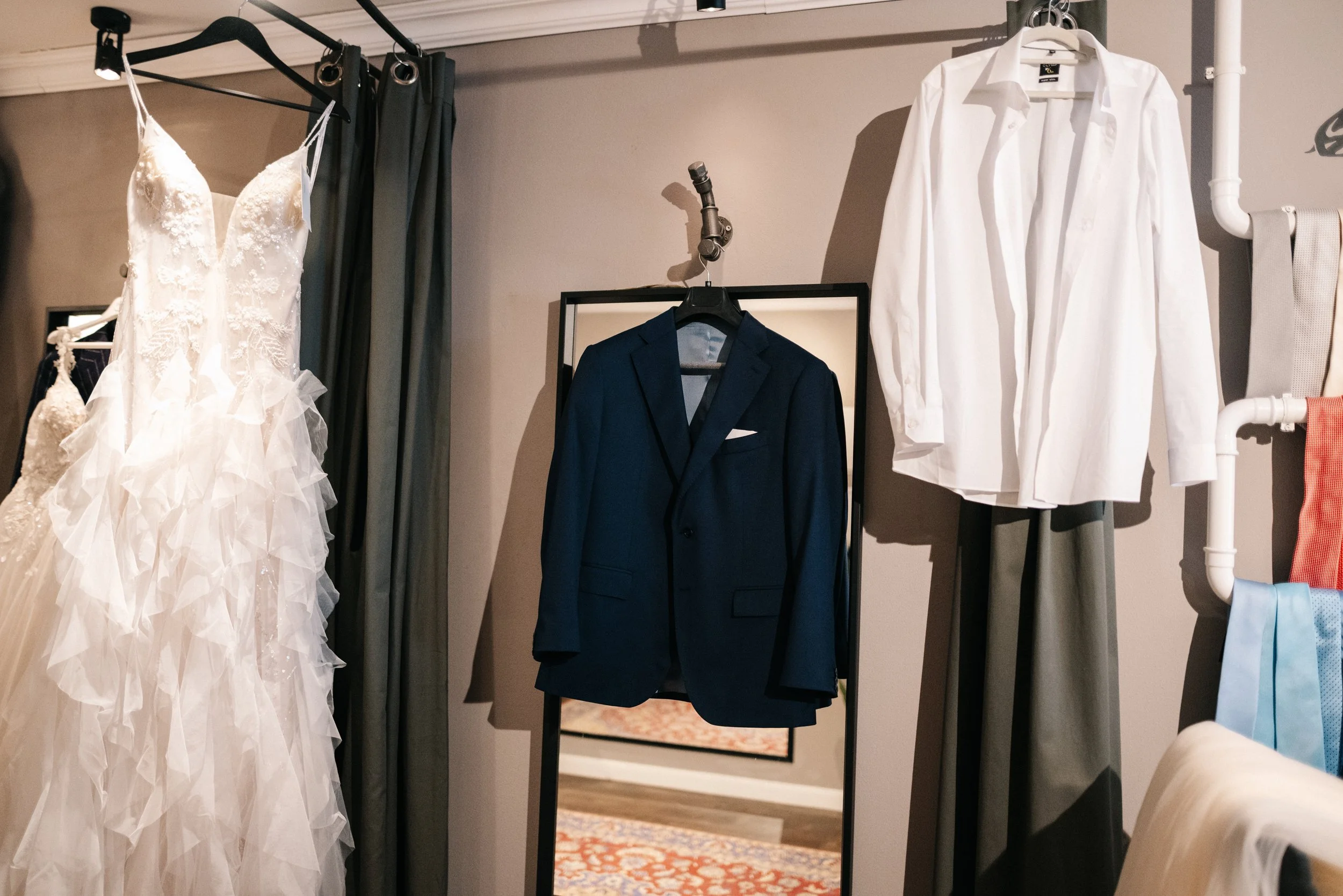 Ein Hochzeitsanzug, ein weißes Hemd, ein weißes Brautkleid mit Spitze und Tüll, und mehrere Stoffe in verschiedenen Farben hängen in einem Ankleidezimmer.