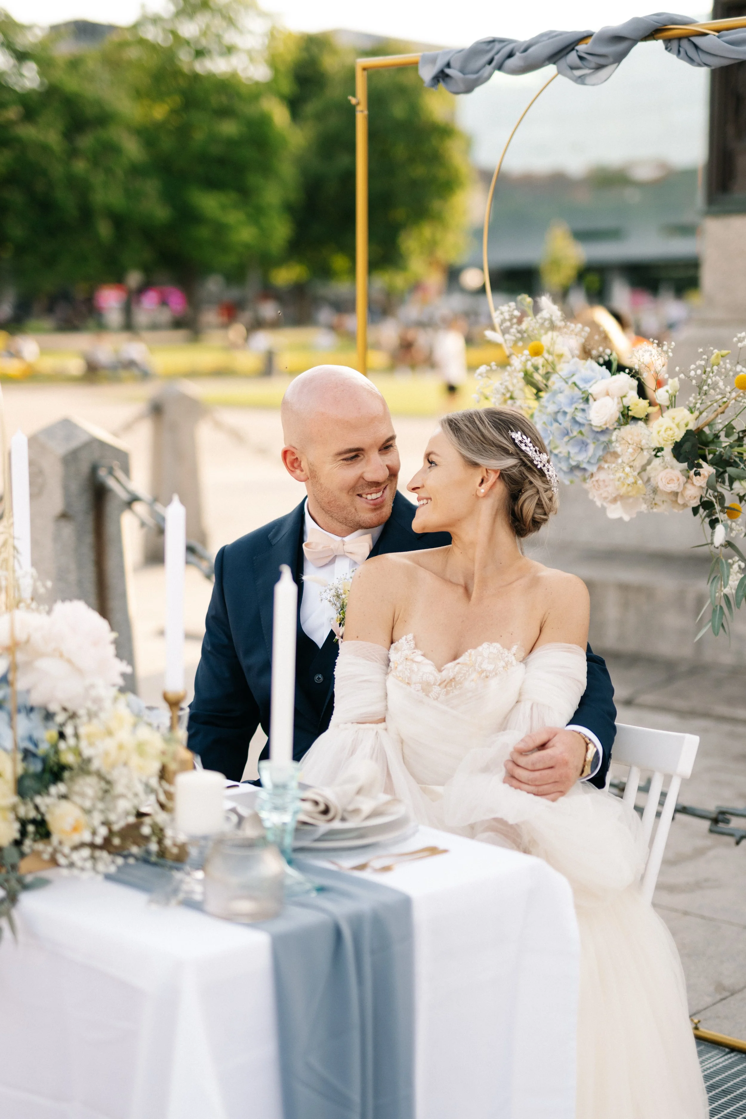 Ein heiratendes Paar sitzt an einem festlich gedeckten Tisch im Freien, umgeben von Blumen, während sie liebevoll in die Augen schauen.