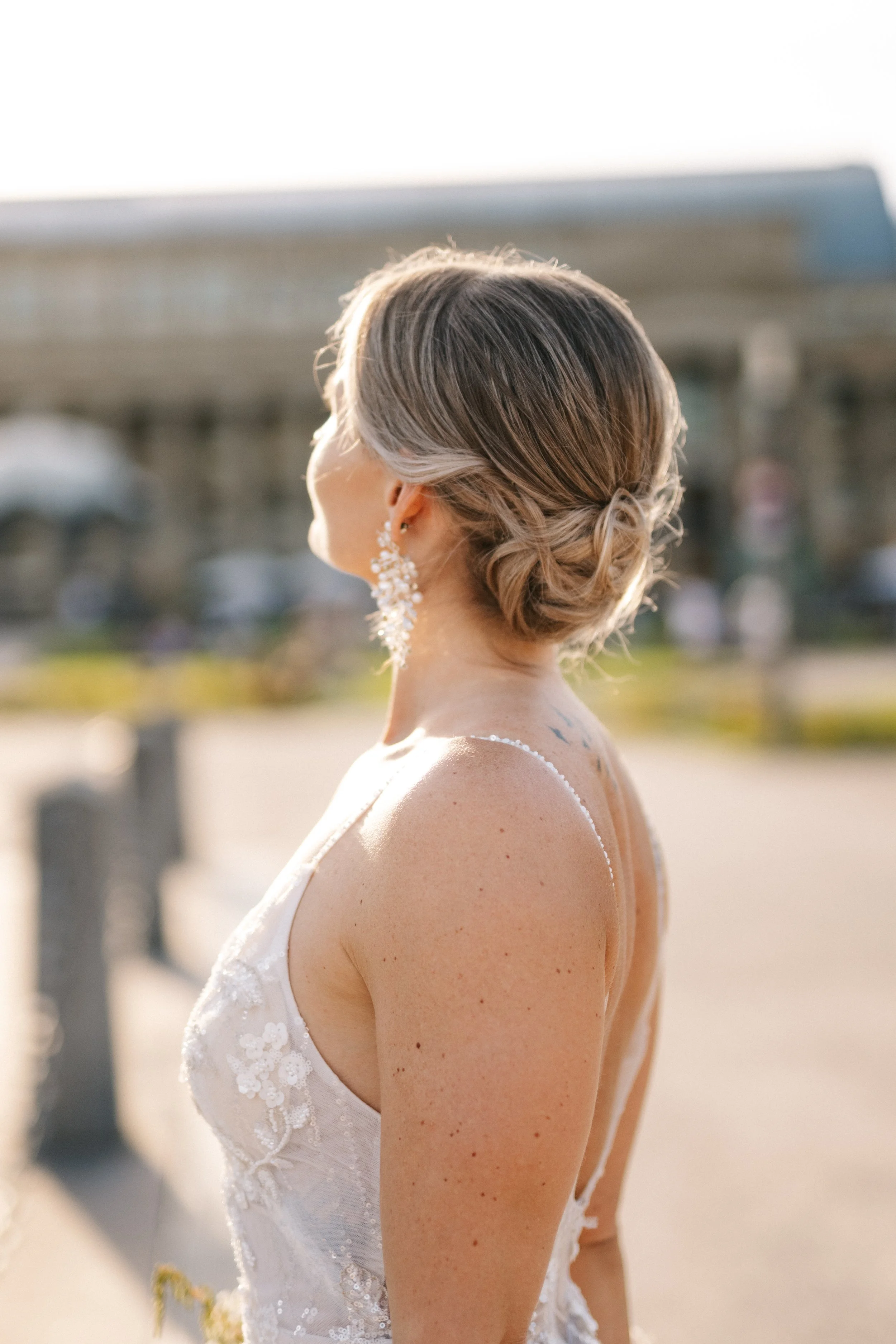 Frau in Brautkleid und eleganten Ohrringen steht im Freien, ihr Rücken ist zur Kamera gerichtet, Sonnenlicht umgibt sie.