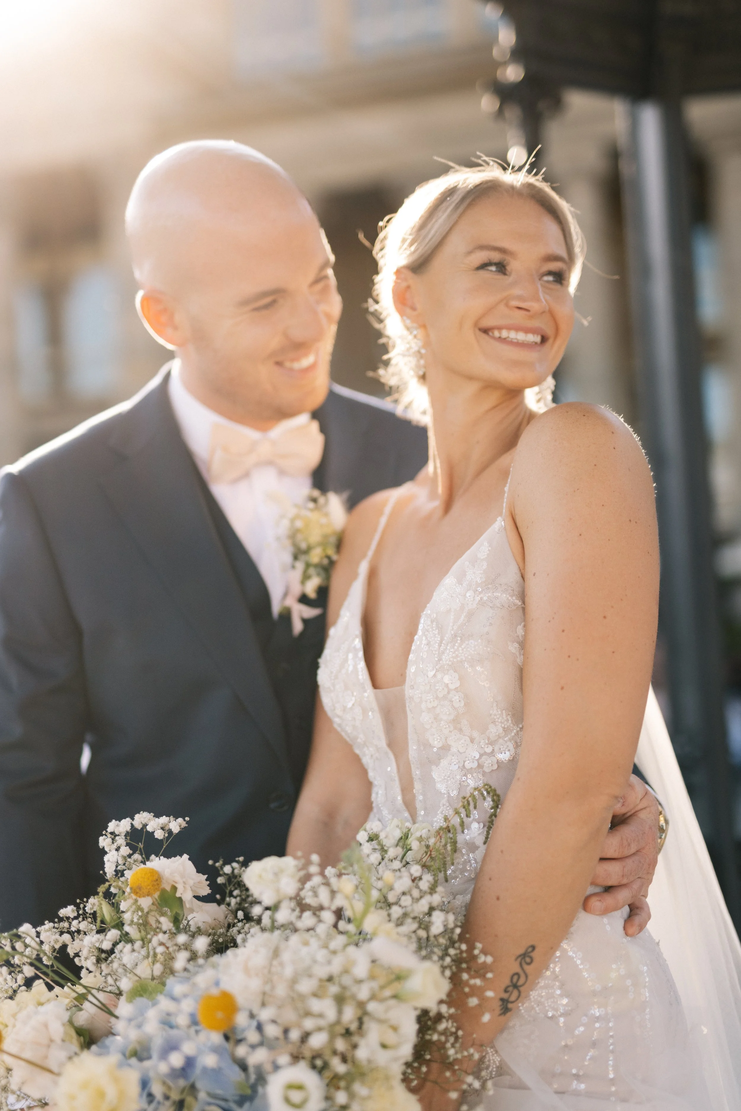 Ein glückliches Brautpaar bei einer Hochzeit im Freien, die Frau trägt ein weißes Hochzeitskleid, der Mann einen schwarzen Anzug mit Fliege, sie hält einen Blumenstrauß, beide lächeln und sind in warmem Sonnenlicht.