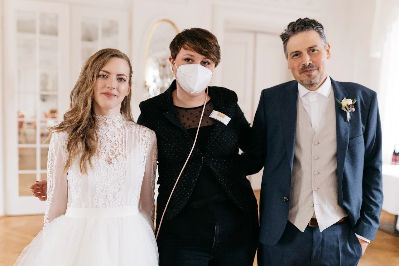 Drei Menschen bei einer Hochzeit, eine Braut in weißem Kleid, ein Bräutigam in Anzug und eine Frau mit Mundschutz in formeller Kleidung.