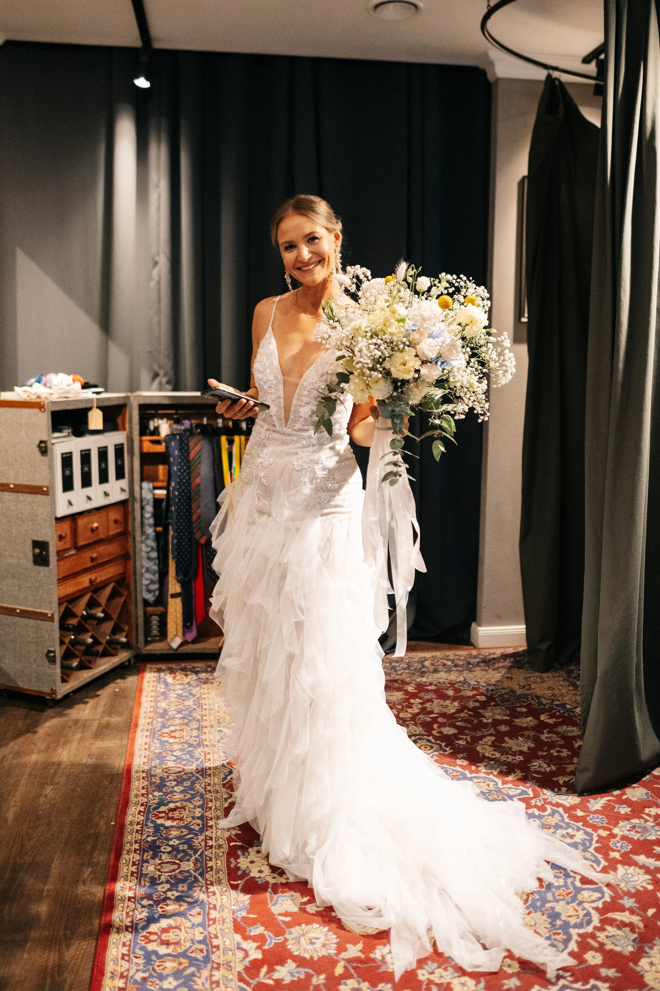 Braut in weißem Hochzeitskleid mit Blumenstrauß, lächelnd, im Inneren eines Raumes mit dunklen Wänden und Teppich, hält ein Handy in einer Hand.