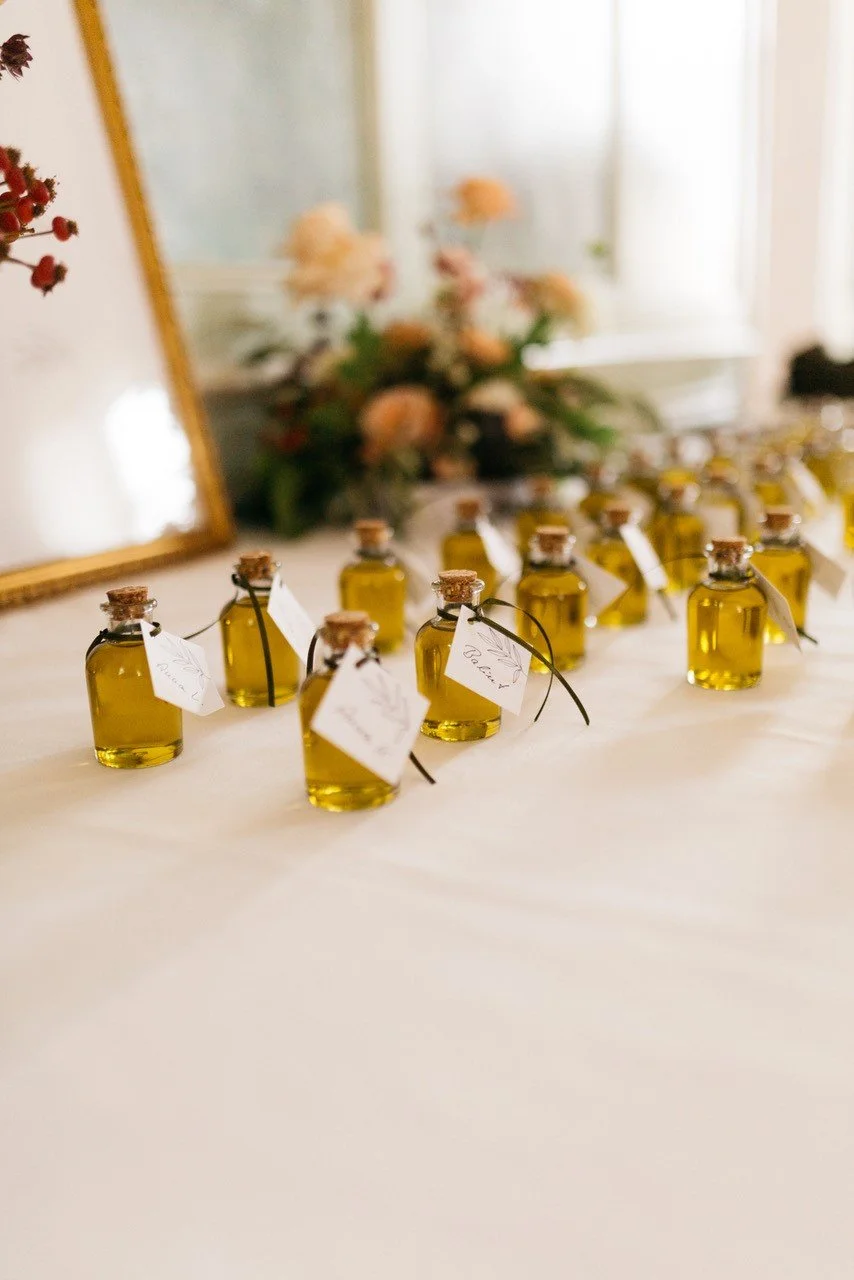 Kleine Flaschen mit gelbem Öl, vermutlich duftendes Öl, mit kleinen Korken und weißen Anhängern auf einem weißen Tisch, im Hintergrund Blumen in einer Vase.