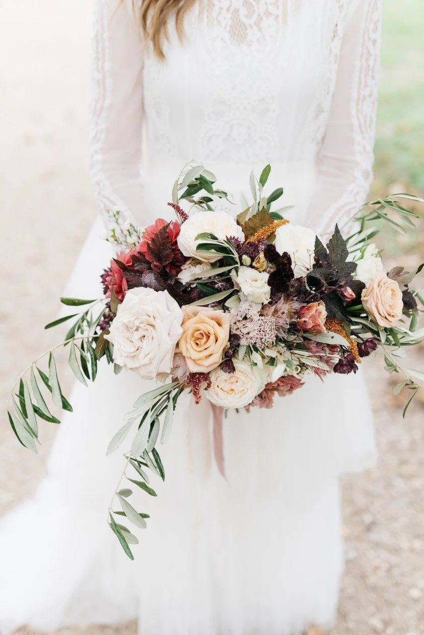 Braut hält einen bunten Blumenstrauß mit weißen, peachfarbenen, roten und dunkelvioletten Blumen, umgeben von grünen Blättern.