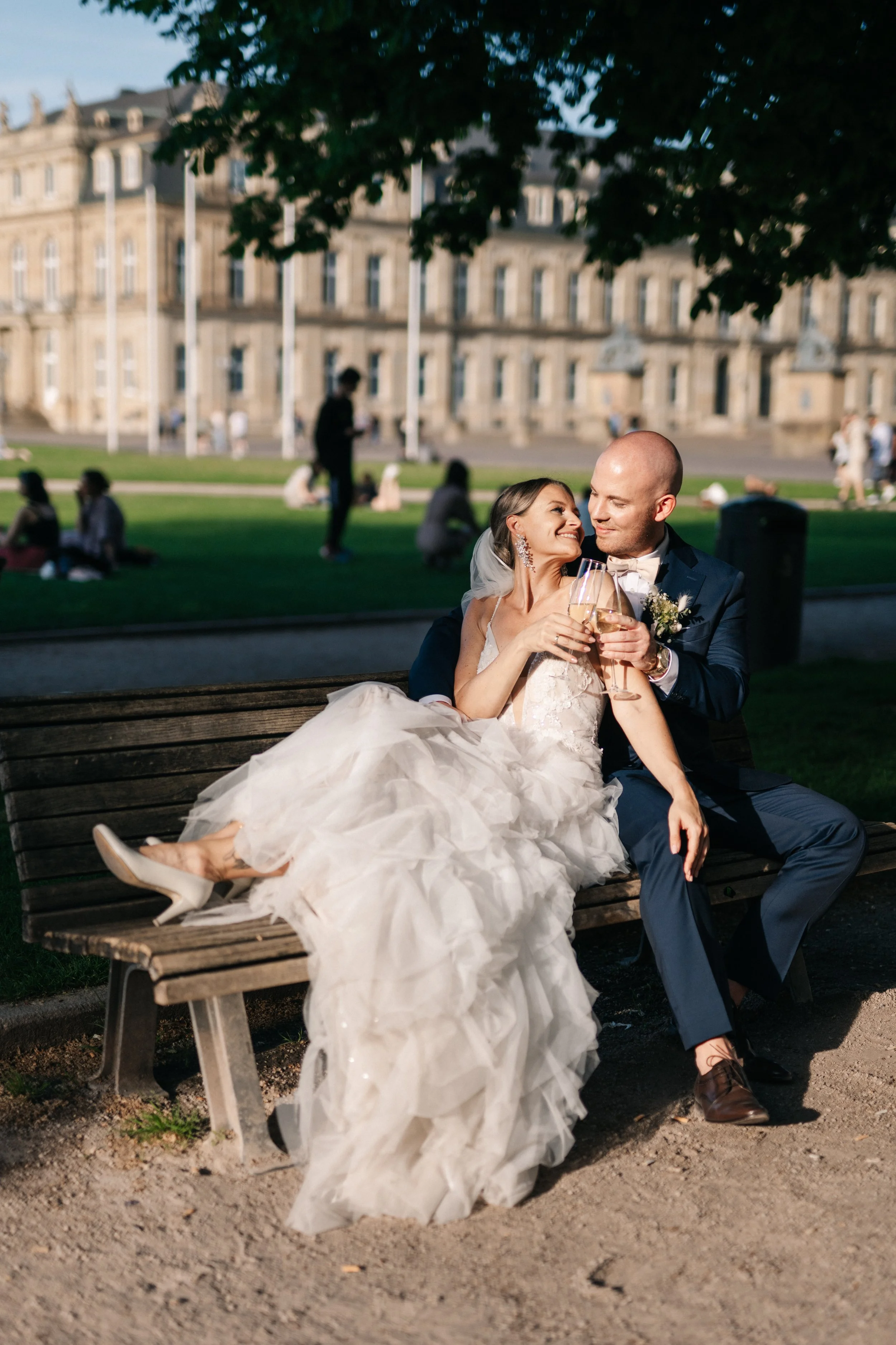 Ein Brautpaar sitzt auf einer Parkbank, trinkt Champagner, glücklich nach ihrer Hochzeit, im Hintergrund ein beeindruckendes historisches Gebäude