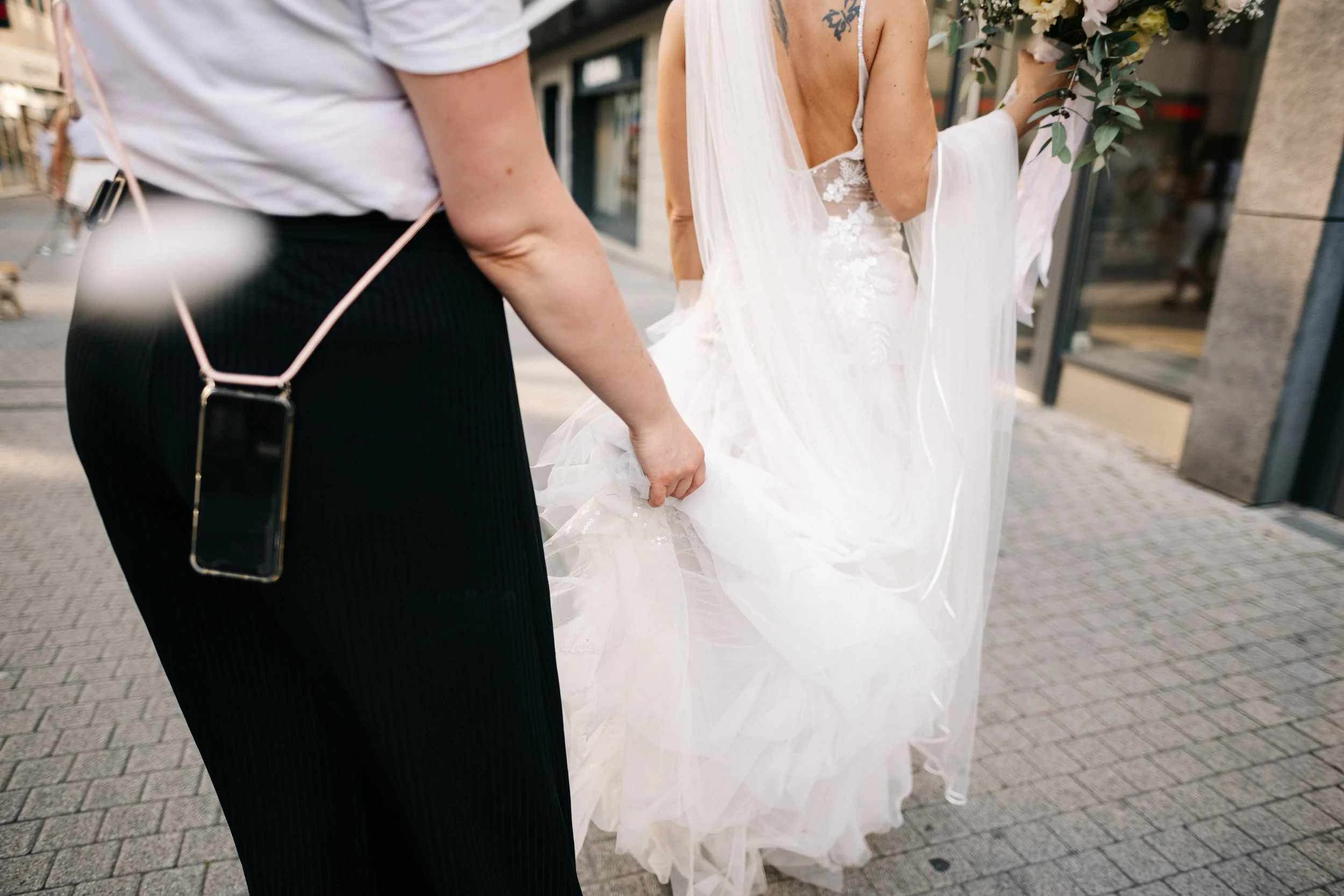 Eine Person in einem weißen Hochzeitskleid hält ein Efeu-Gesteck, während eine andere Person die Brautkleid hochhebt, auf einem Bürgersteig vor einem Geschäft