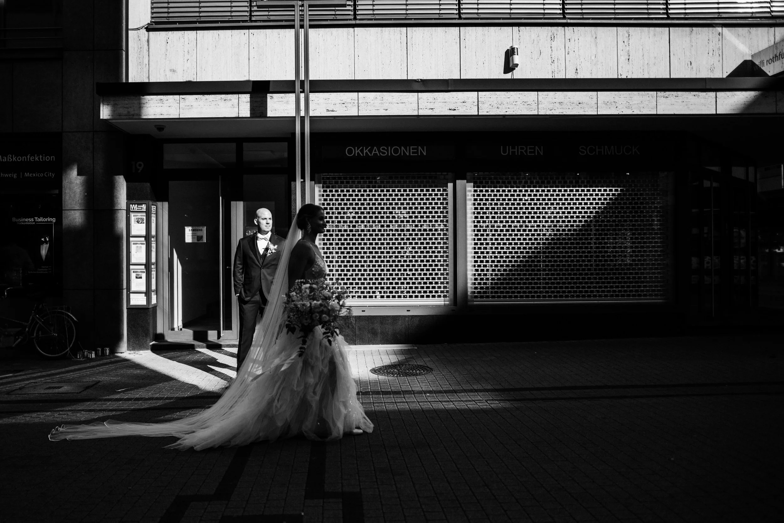 Schwarzweiße Aufnahme eines Brautpaares, das Seite an Seite an einer urbanen Straße steht, mit Schatten und Licht. Die Braut trägt ein langes Brautkleid mit Schleier, während der Bräutigam einen Anzug trägt.