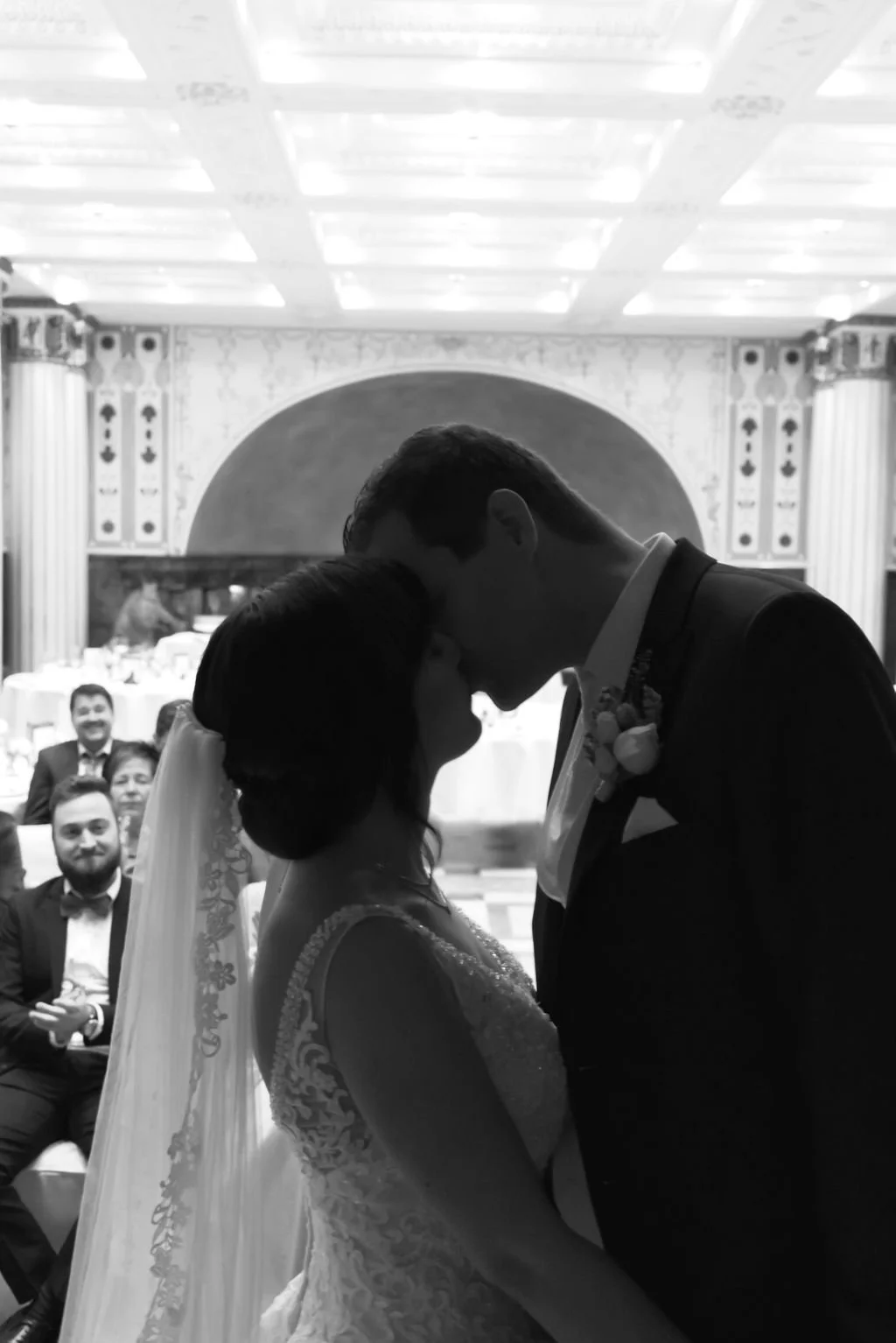 Braut und Bräutigam umarmen sich bei ihrer Hochzeitszeremonie, im Hintergrund sitzen Gäste im festlichen Saal.