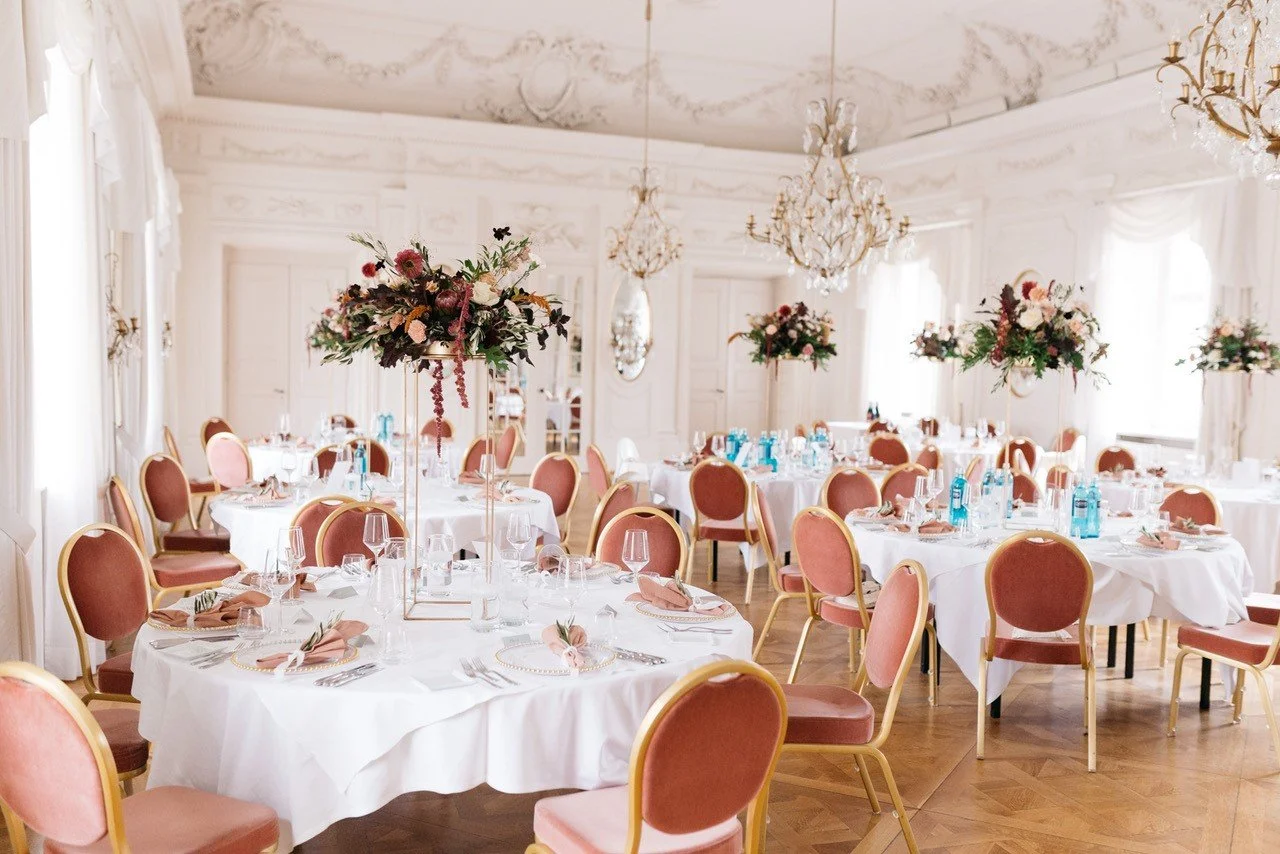 Elegant festlich gedeckter Saal mit runden Tischen, blumigen Mittelstücken, Wasserflaschen, Gläsern, Besteck und beige-rosa Stühlen, für eine Veranstaltung oder Hochzeit.