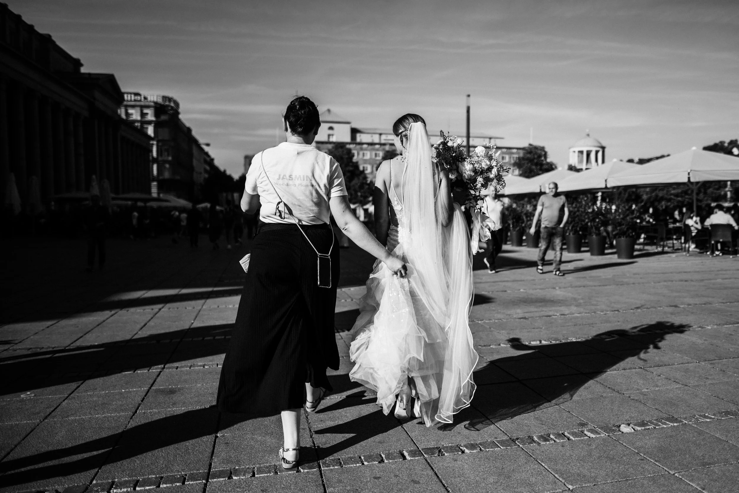 Eine Frau in einem Hochzeitskleid mit Schleier hält einen großen Brautstrauß, während sie von einer Person in schwarzer Hose und weißem T-Shirt begleitet wird, in einer urbanen Umgebung bei Sonnenuntergang.